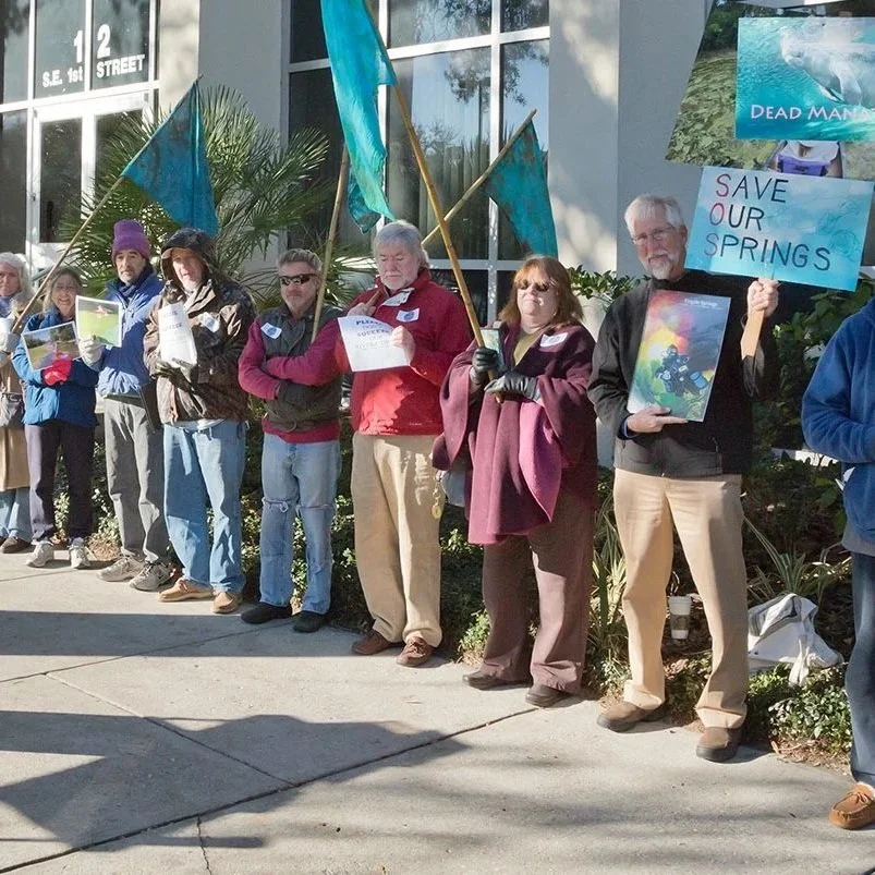 A speaker addresses a group of environmental advocates gathered outside a building during a clean water rally, while participants hold signs and images supporting the protection of Florida’s springs.