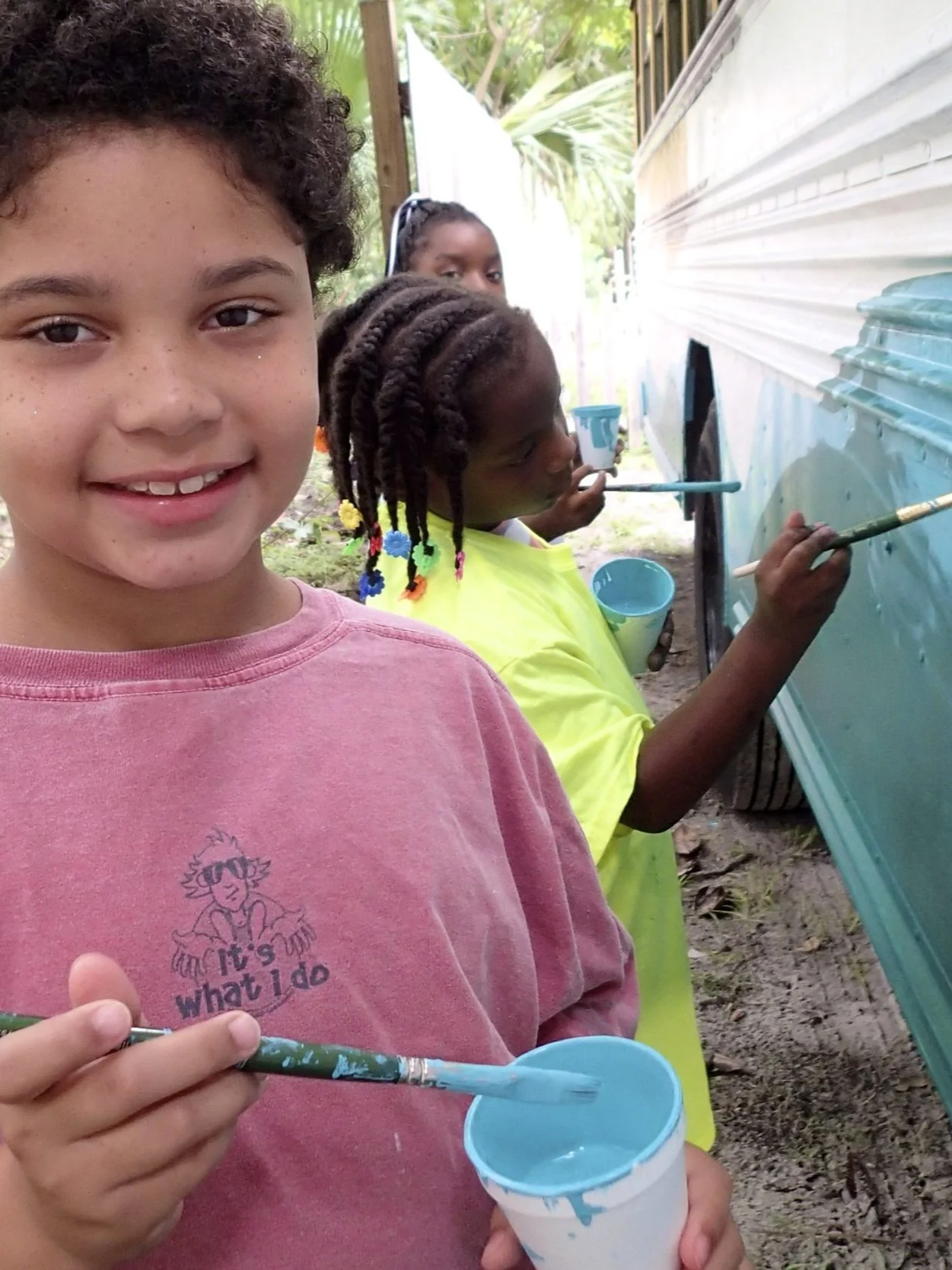 Child painting the side of a bus with a brush and blue paint during a youth art initiative.