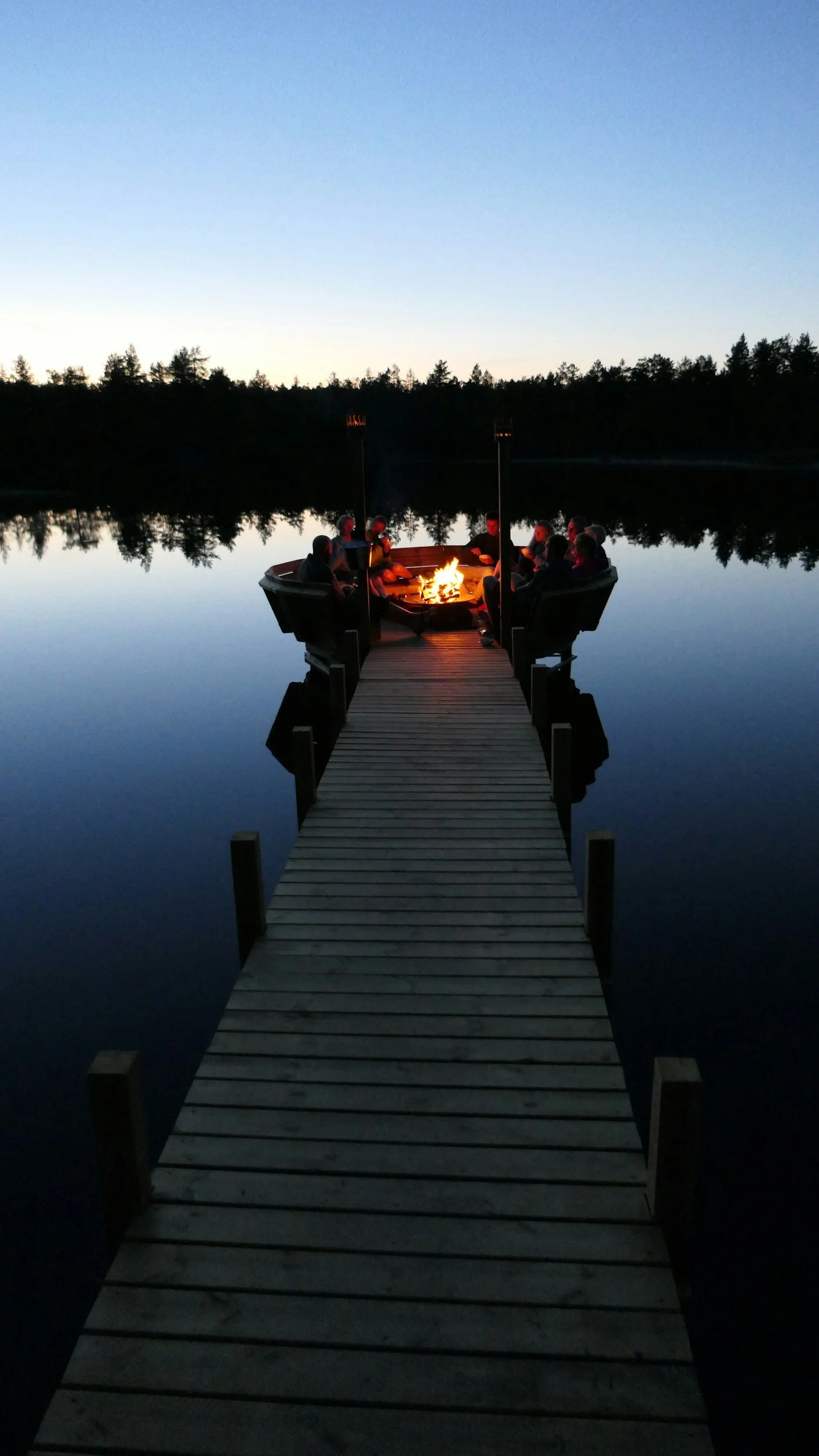 Tomorrow house retreat photo featuring a group of sitting around a fire on a boat at the end of a dock on a calm lake during dusk.