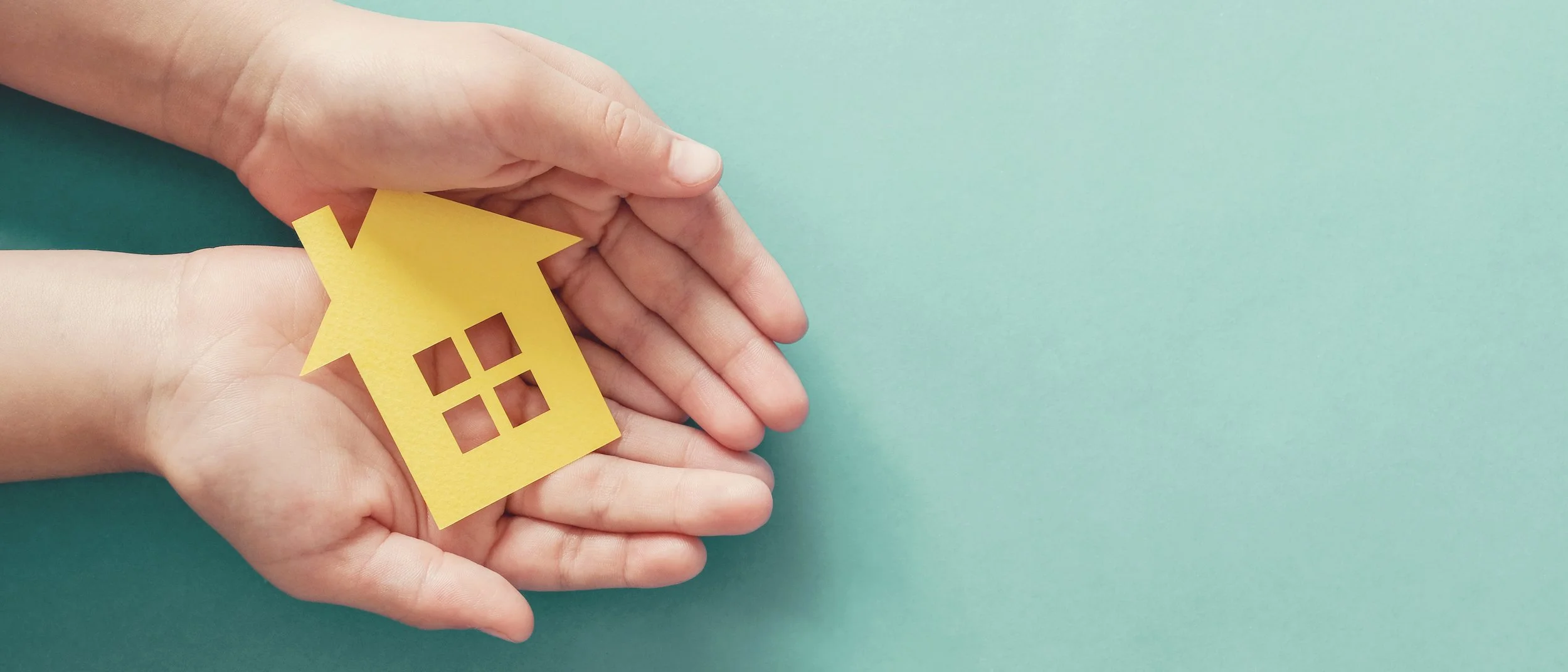 Two hands holding a small yellow paper house cutout with window and roof details on a light green background.