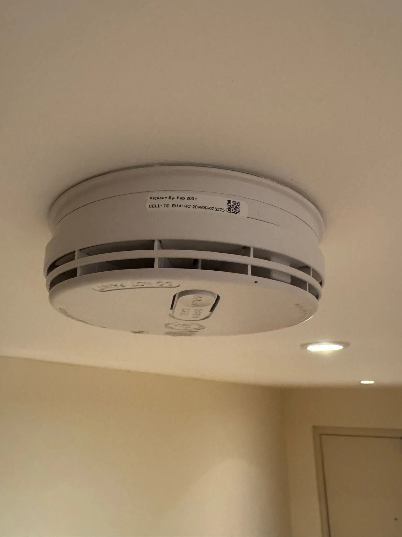 Ceiling smoke detector mounted on the ceiling, with a beige wall and some recessed lights in the background.