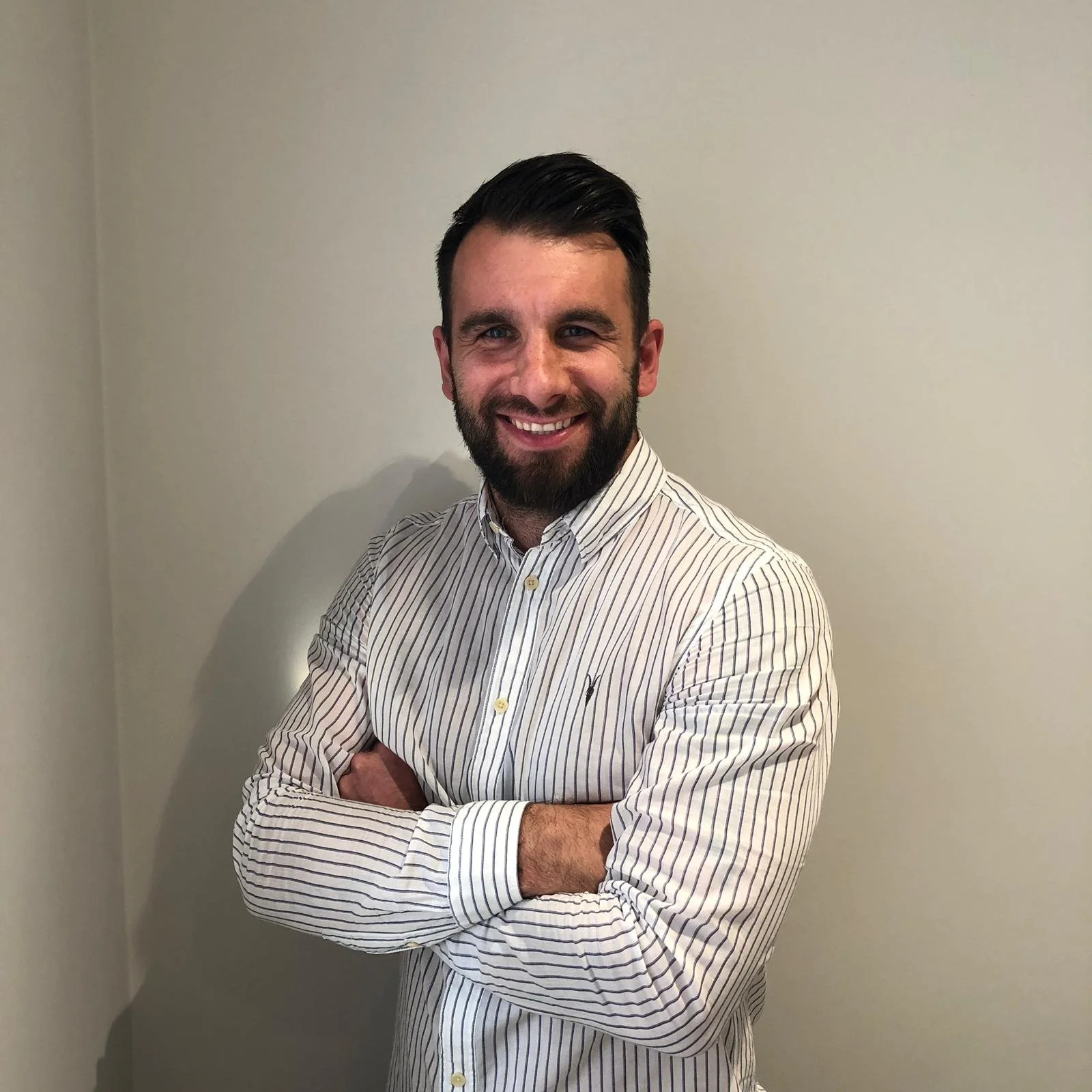 A man with dark hair and a beard, smiling, standing with arms crossed in front of a plain cream wall, wearing a white and gray striped button-up shirt.