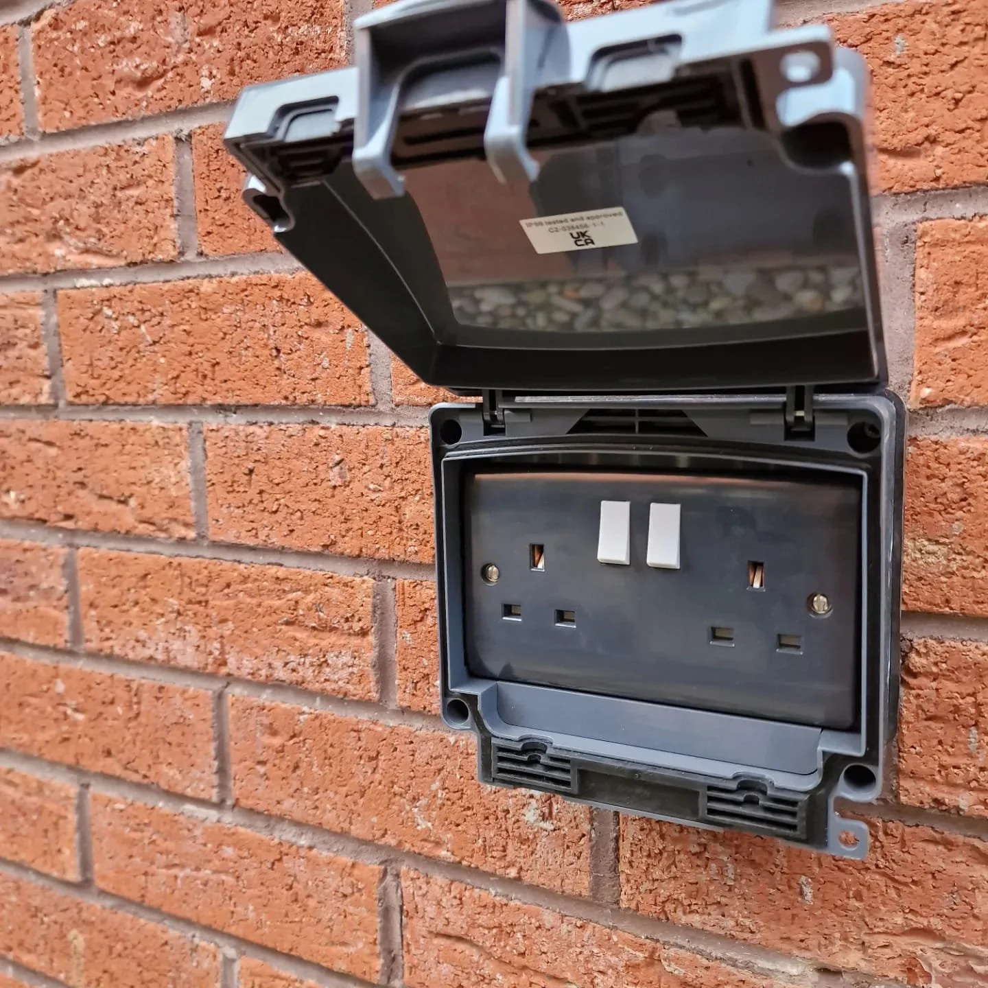 Empty outdoor electrical outlet with a protective cover mounted on a brick wall.