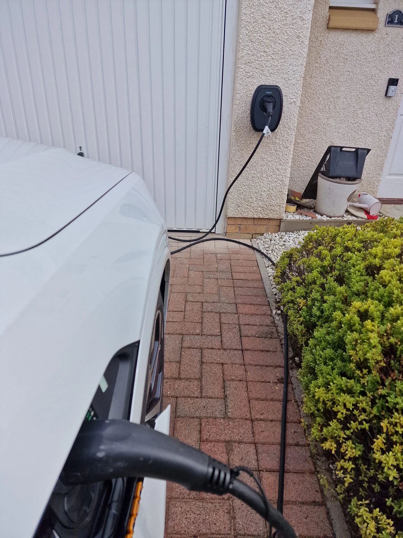 Electric vehicle charging cable plugged into white car near garage door, with a bush and garden tools nearby.