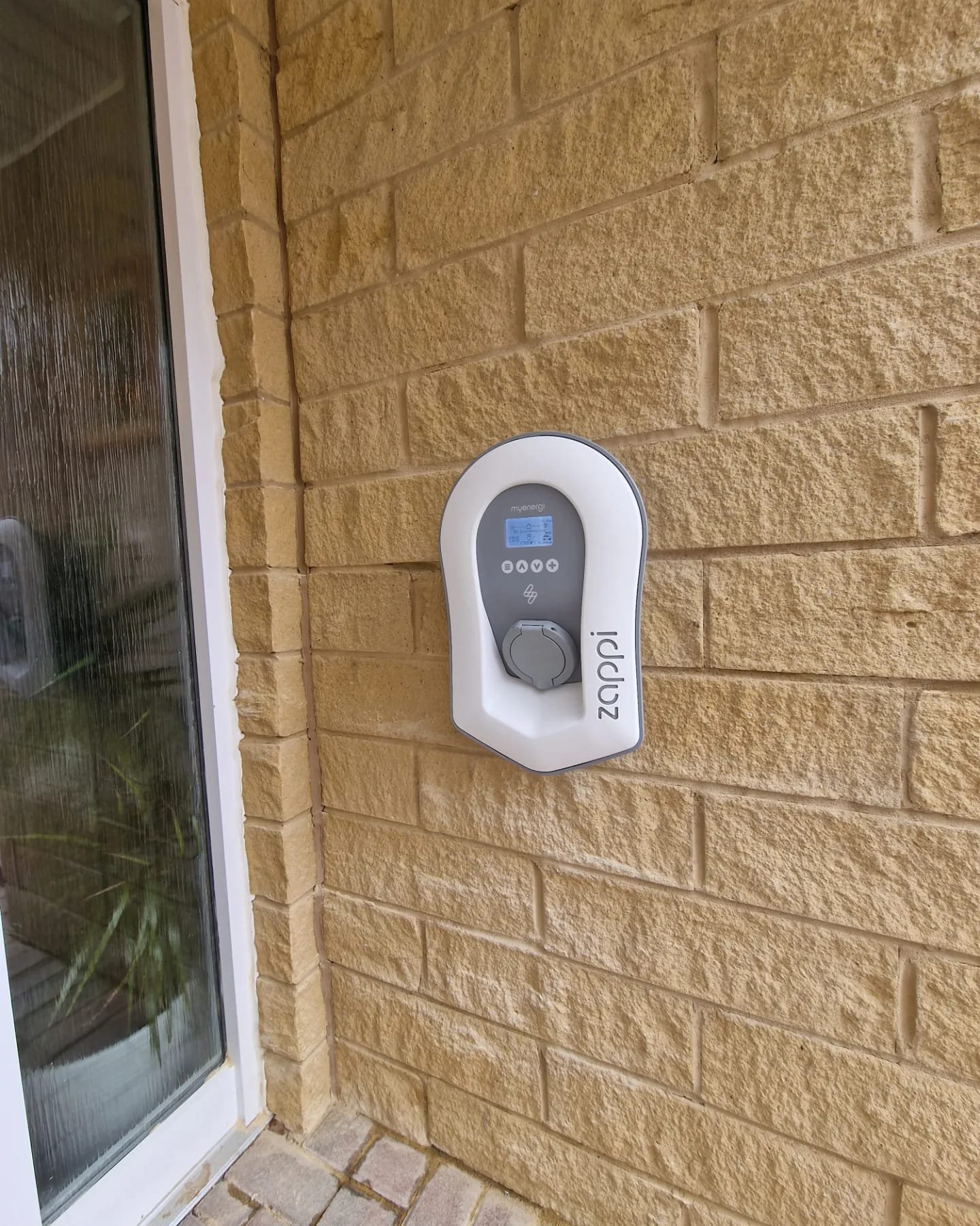 A wall-mounted electric car charging station on a brick wall next to a glass door.