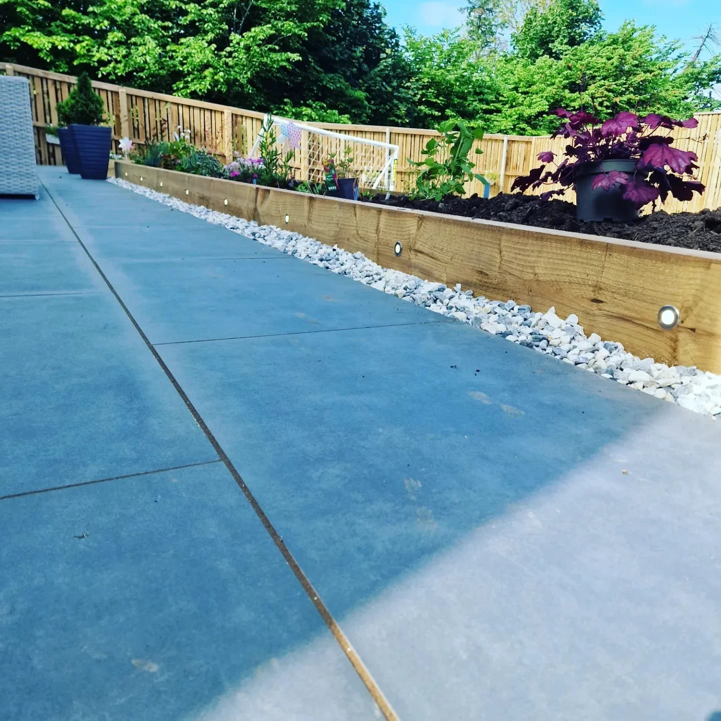 Close-up of a backyard garden bed with colorful flowers and plants, wooden edge, white pebbles lining the bed, and a green fence with trees in the background.