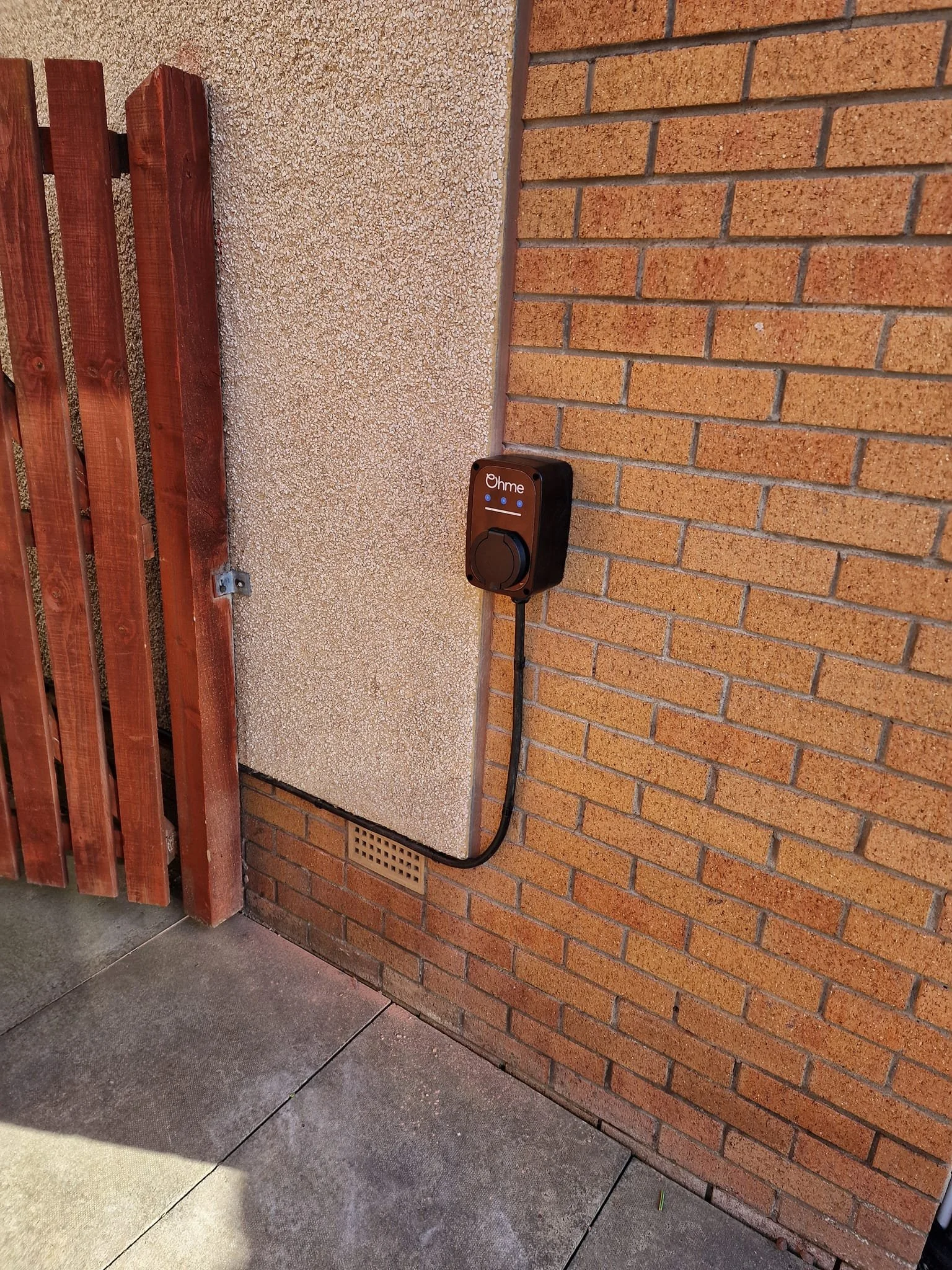 An electric vehicle charger mounted on a brick and stucco exterior wall of a building, with a black cable attached.