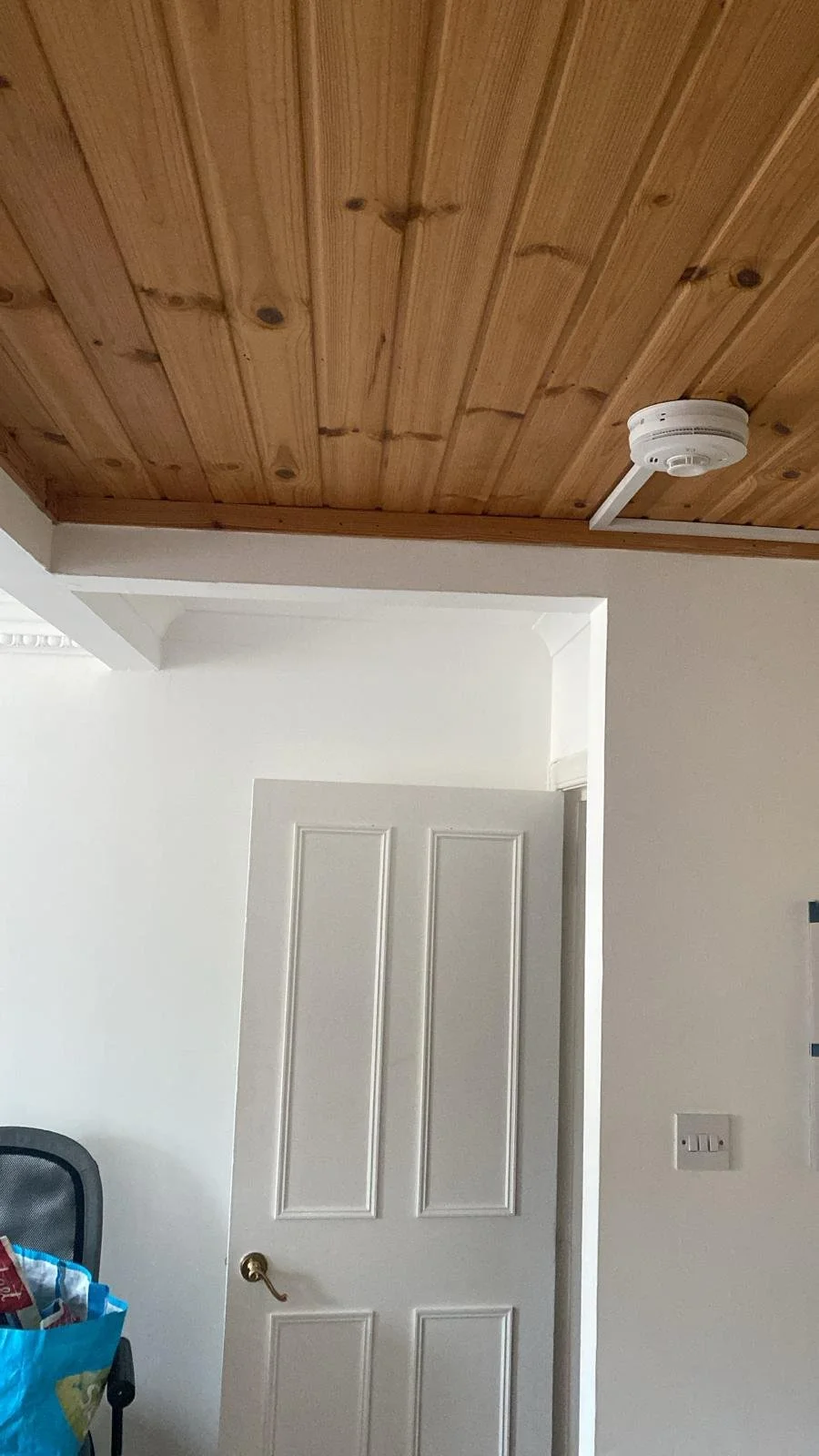 Interior view of a room showing a wooden ceiling, a white door slightly ajar, a wall with a light switch, and part of an office chair with a blue bag on it.