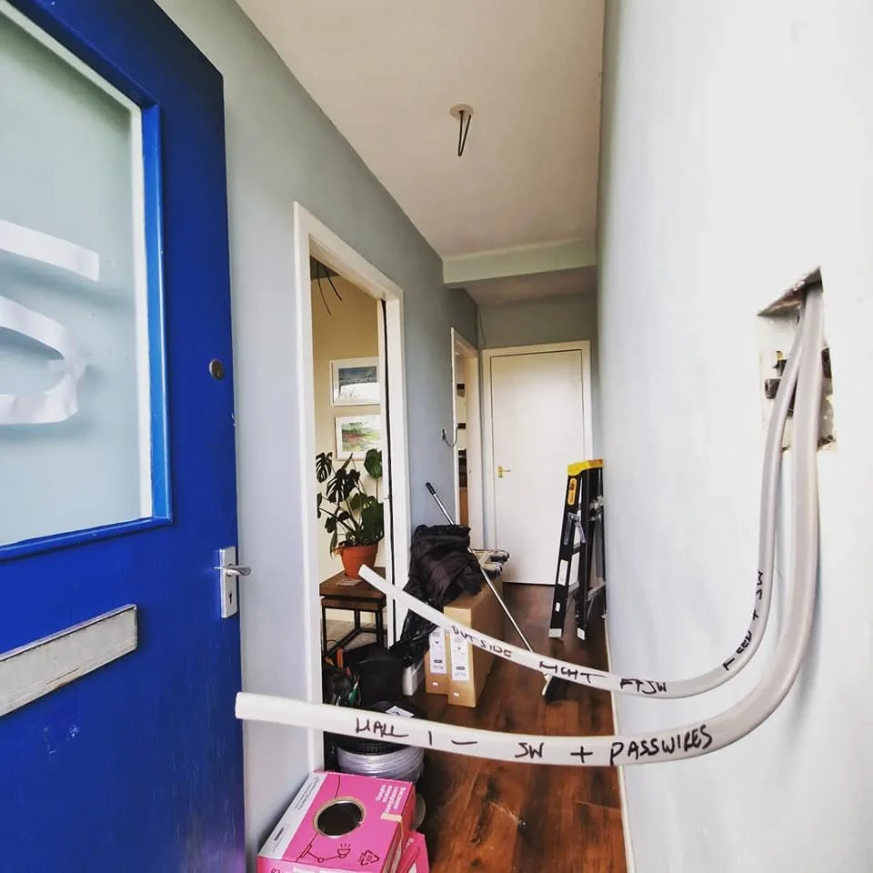 Indoor hallway with construction tools and materials, including a white cable with black writing, a yellow and black step ladder, boxes, and a potted plant, leading to a door at the end.