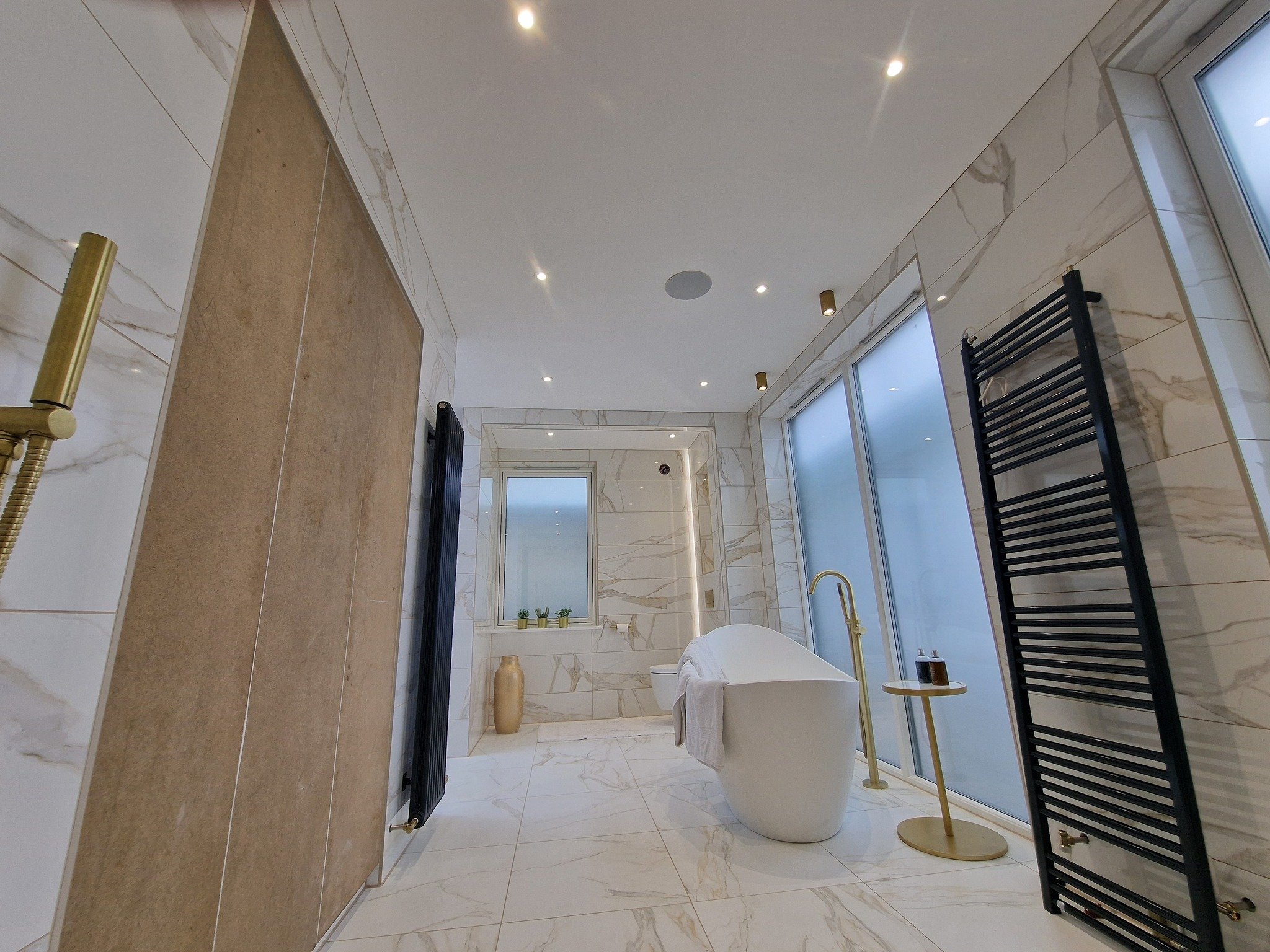 Luxurious bathroom with marble tiles, a freestanding bathtub with a gold faucet, black towel racks, large frosted windows, and decorative vases.