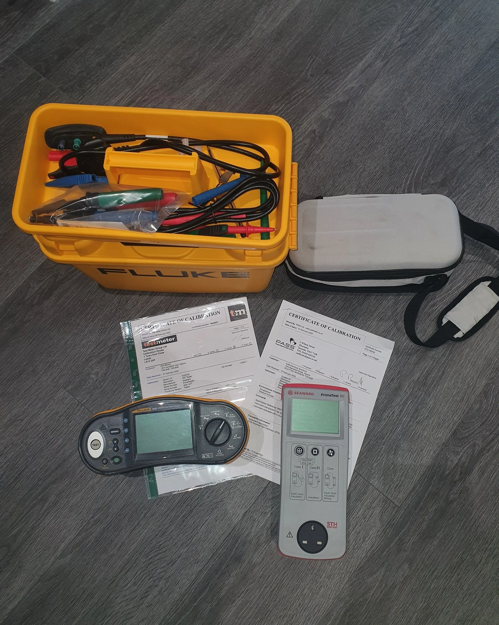 Electrical testing equipment including a Fluke multimeter, a Seaward PrimeTest tester, calibration certificates, and a yellow tool box with wires and probes, on a gray wooden surface.