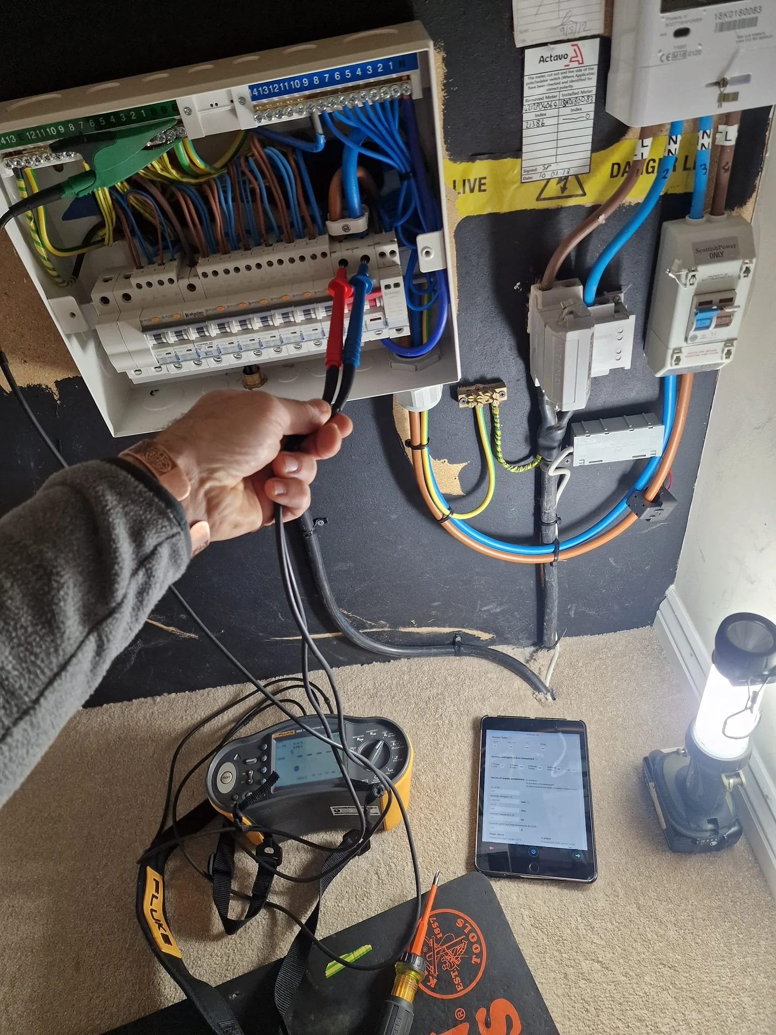 A person testing electrical wiring in an electrical panel with a multimeter. The panel has multiple colored wires and circuit breakers. There is a flashlight, a tablet, and a tool on the carpeted floor.