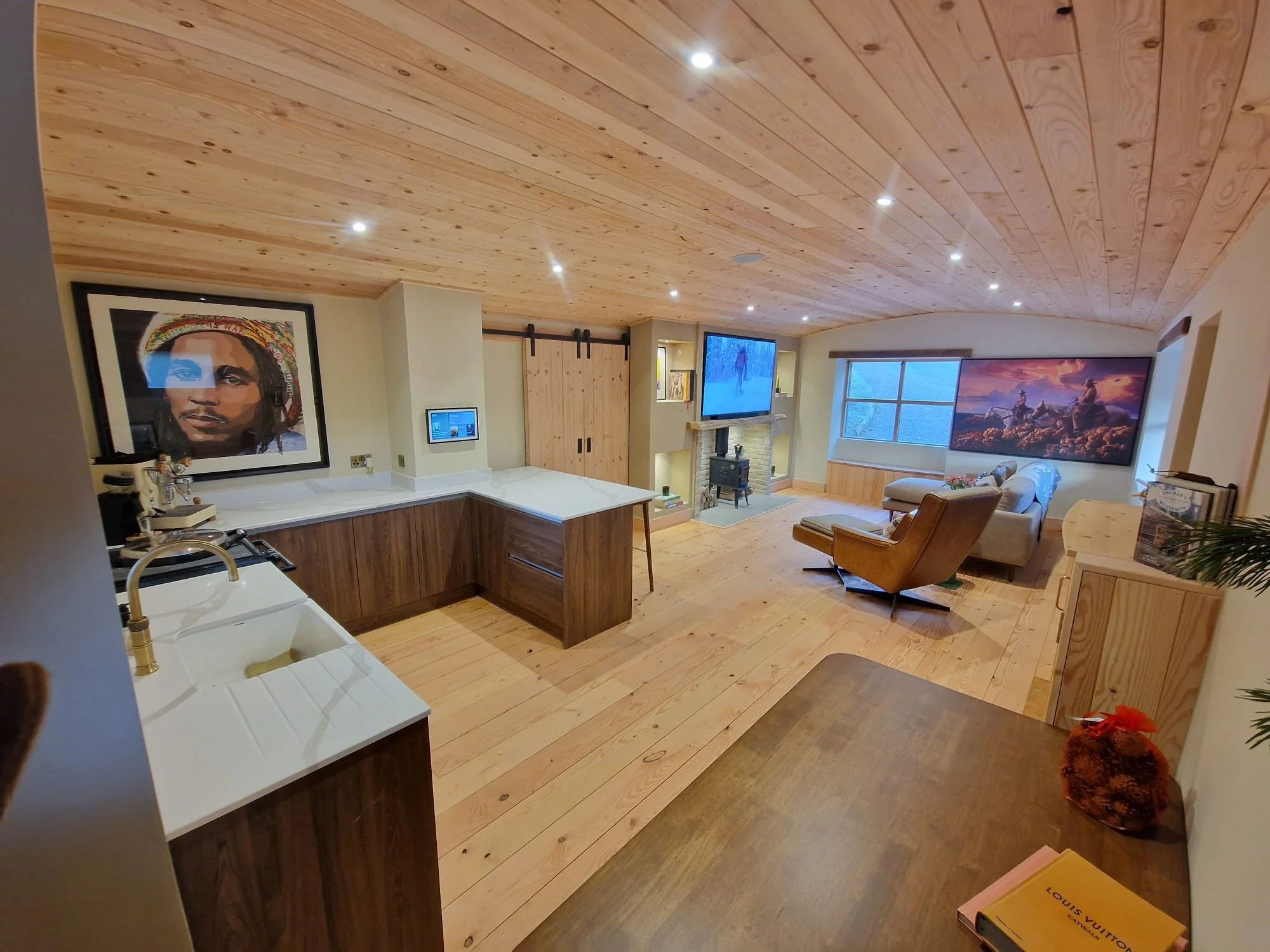 Living room with wooden ceiling and floors, two large windows, a fireplace with a chimney, a flat-screen TV, a beige couch, a brown armchair, and artwork on the walls.