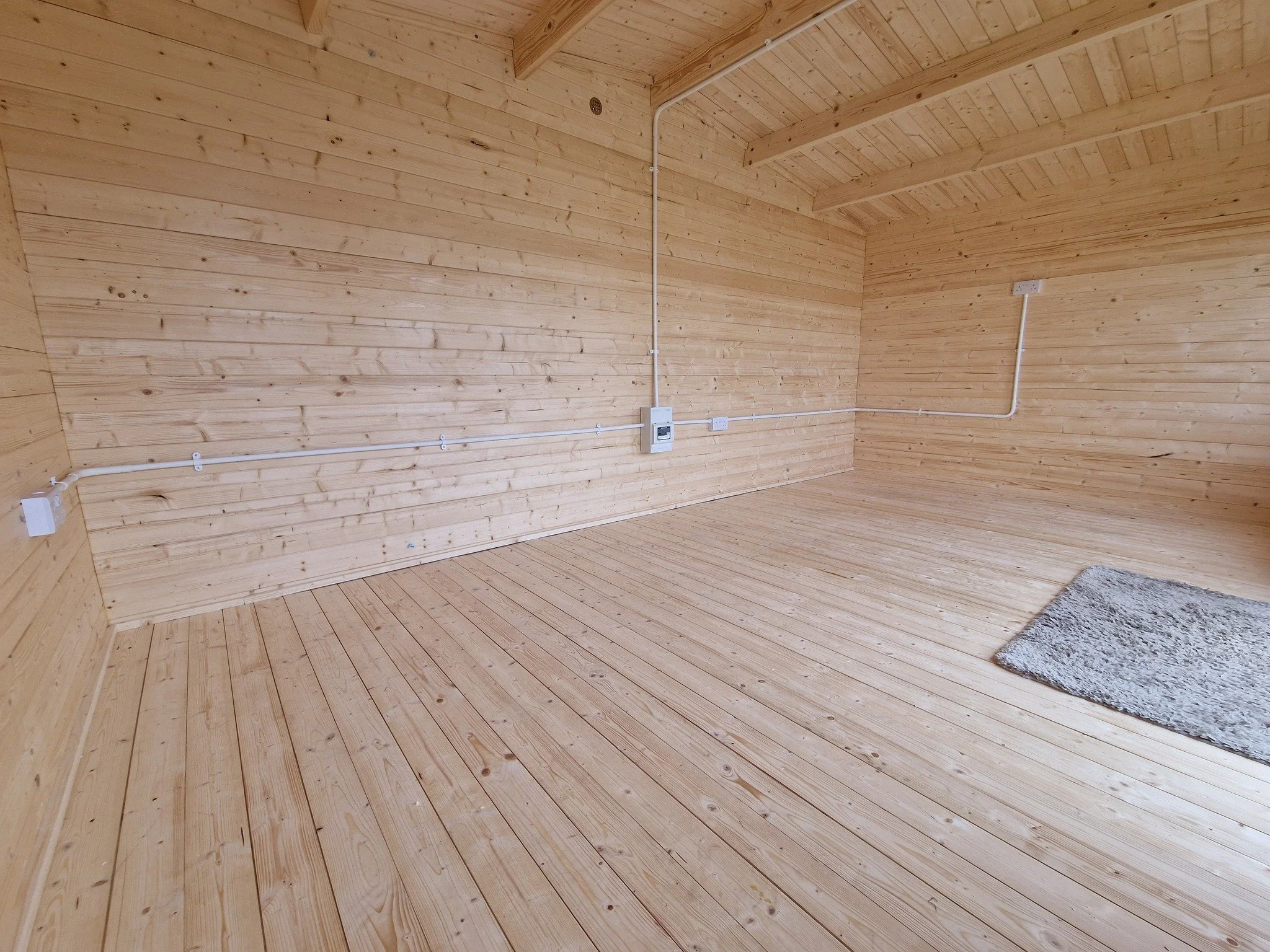 Empty room with wooden walls, floor, and ceiling, with visible electrical outlets and a small gray rug on the floor.