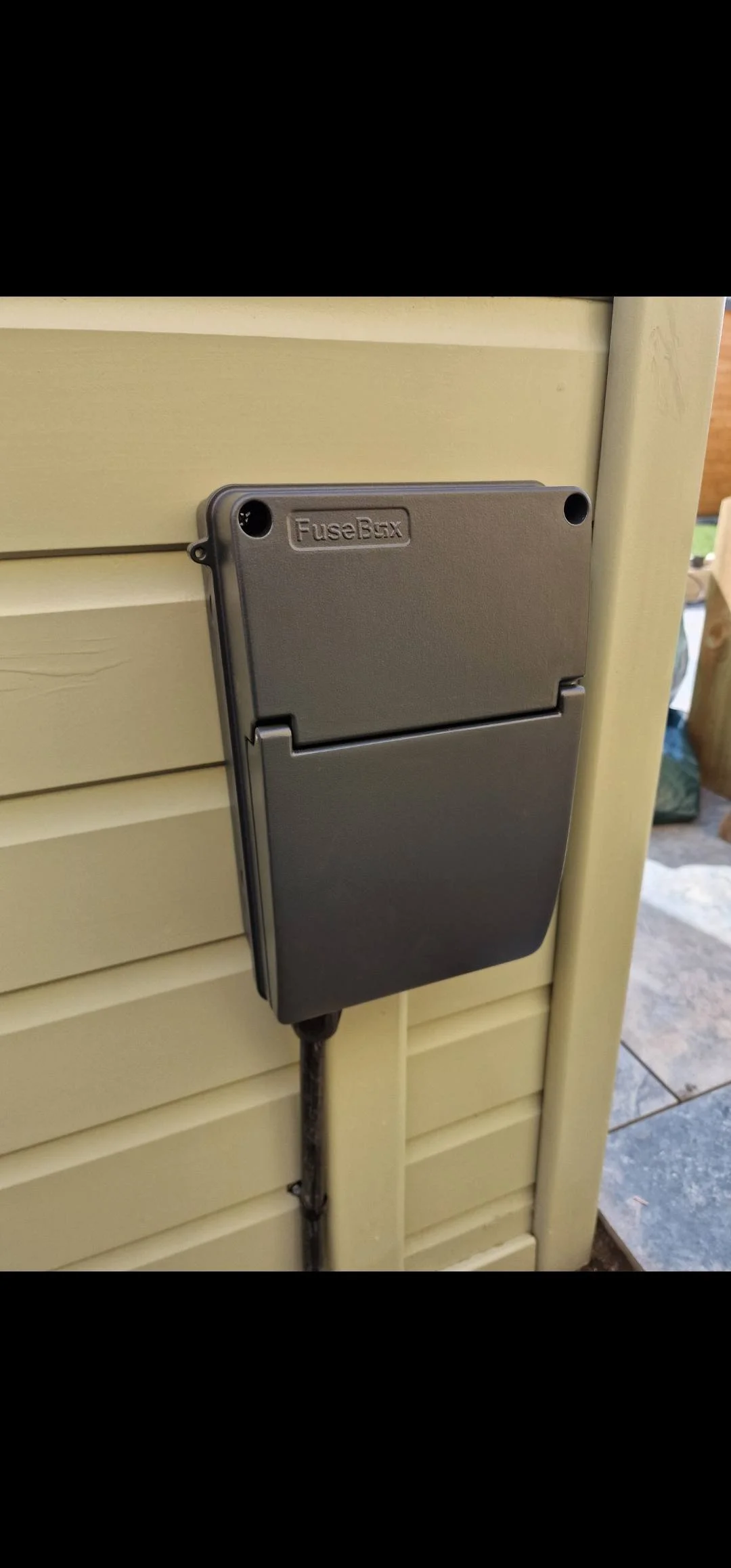 A gray electrical box labeled FuseBox mounted on a beige wooden wall with a black cable hanging down.