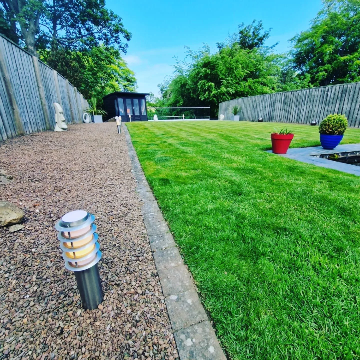 Well-maintained backyard with green grass, decorative pots, a wooden fence, small garden shed, and landscape lighting.