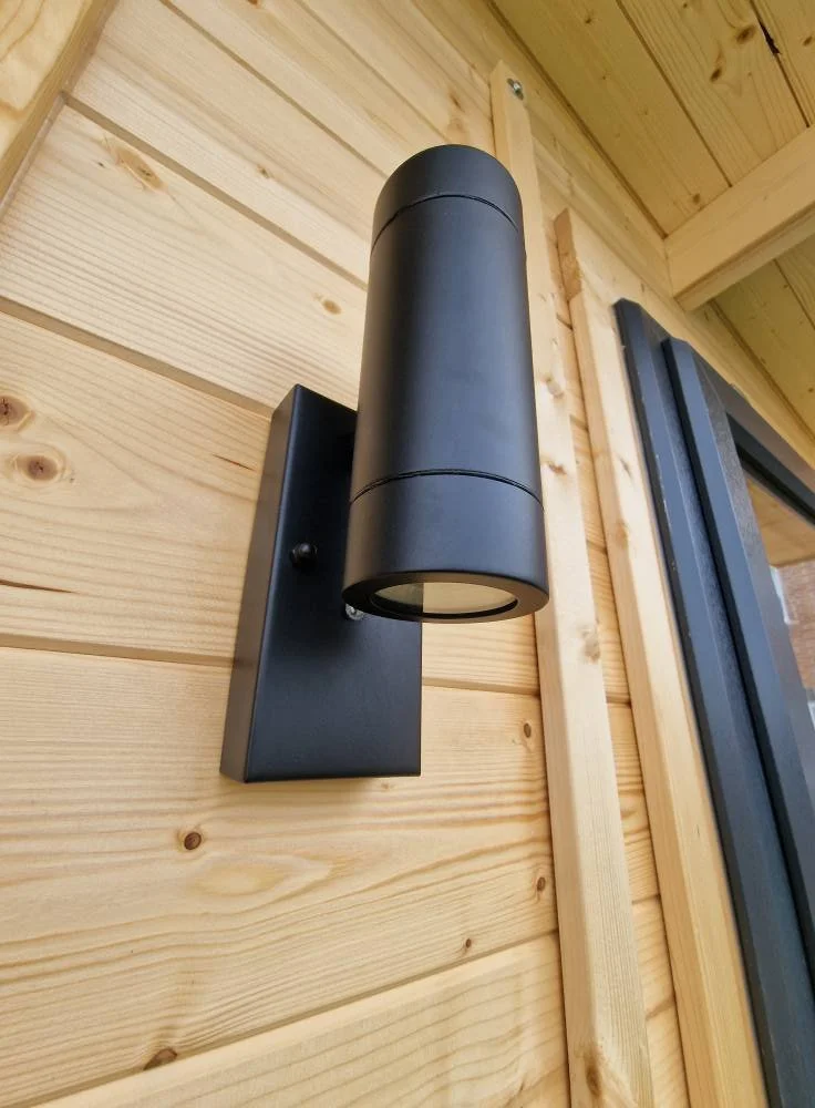 Black outdoor wall sconce light fixture mounted on light wood-paneled wall near window.