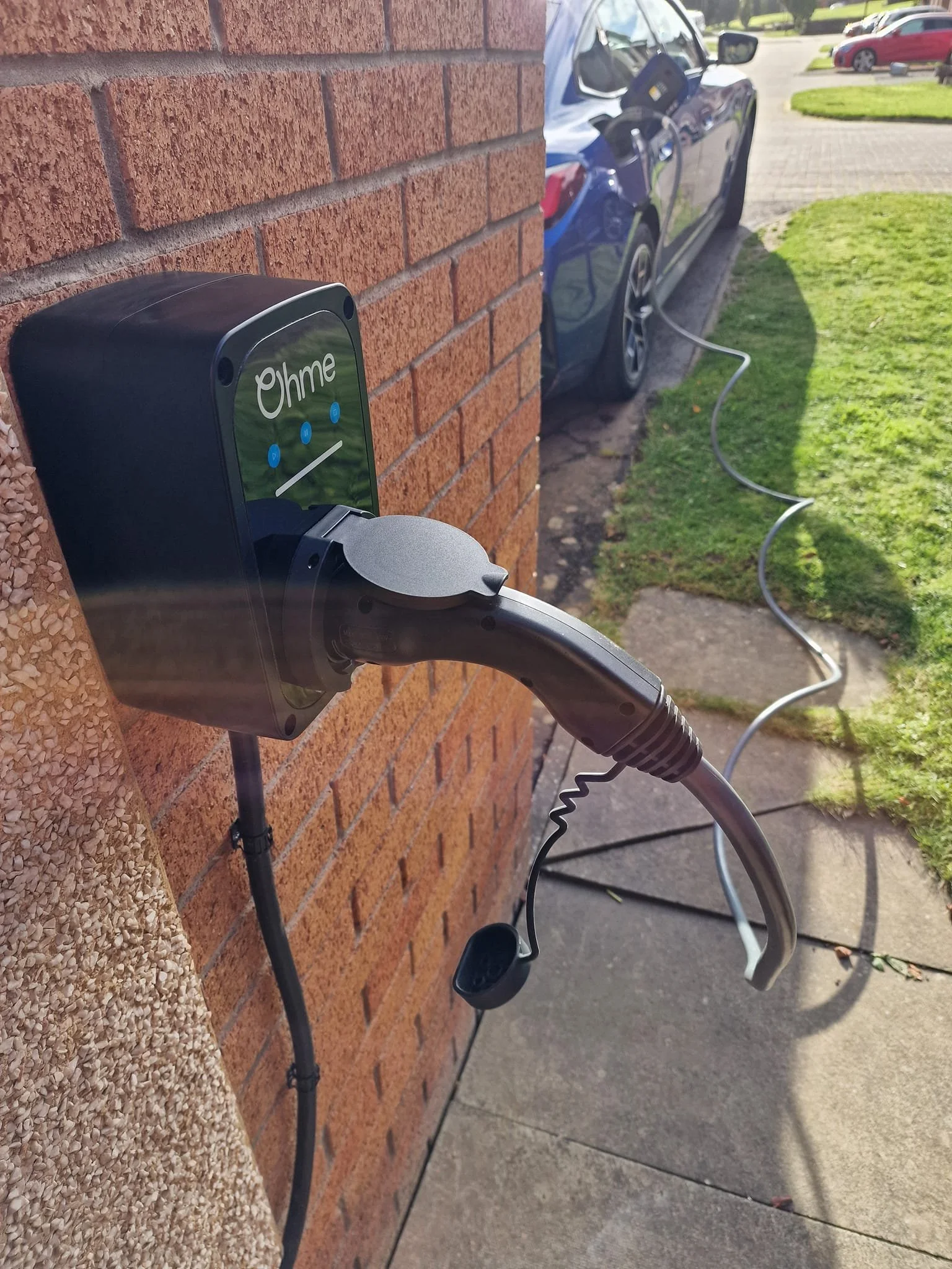 Electric vehicle charging station attached to a brick wall, with a charging cable connected to a blue electric car parked outside on a sidewalk.