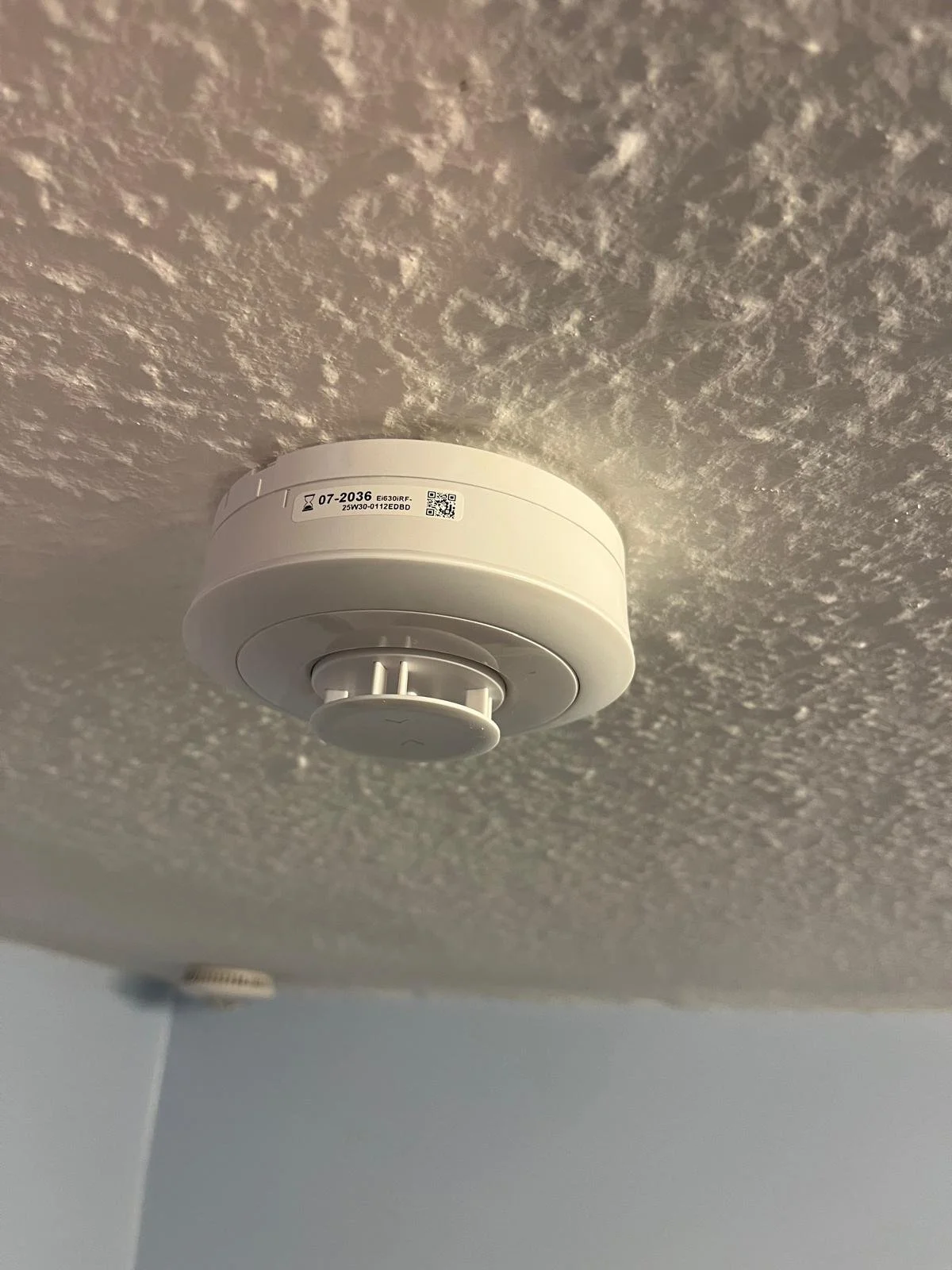 Smoke detector mounted on ceiling with textured ceiling surface in the background.
