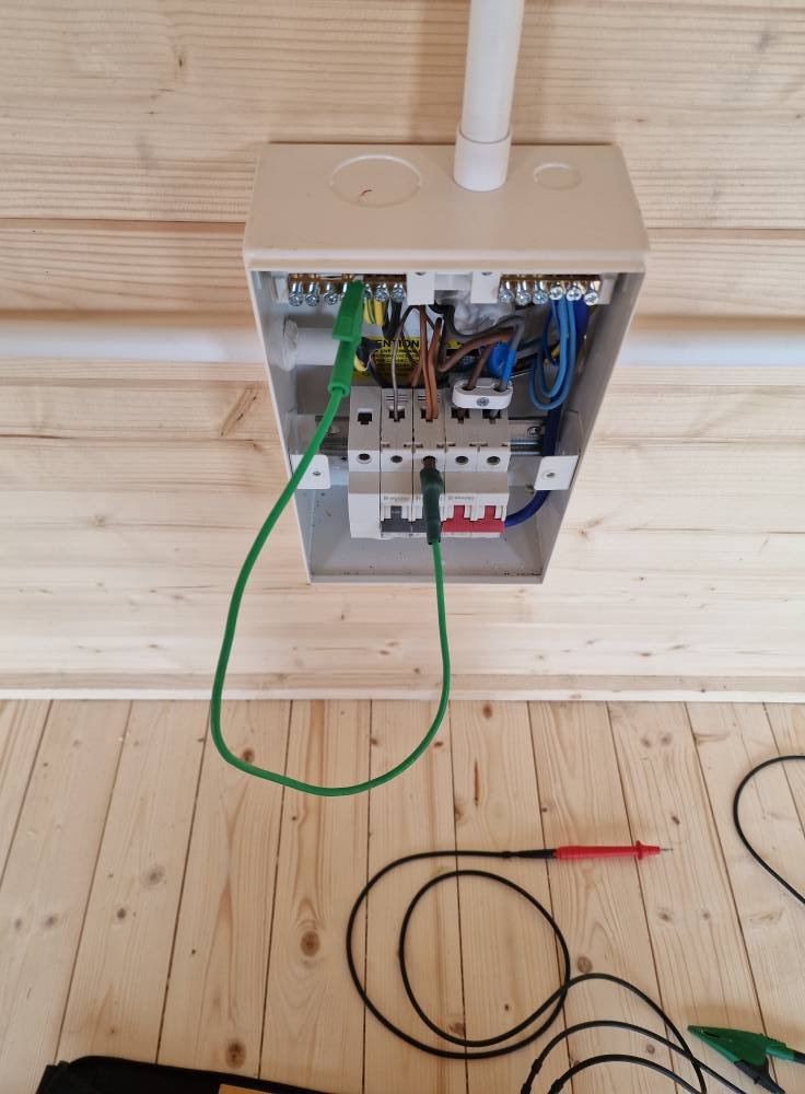 Open electrical junction box with wires connected inside, a green wire connected to the outside, and testing tools on the wooden floor below.