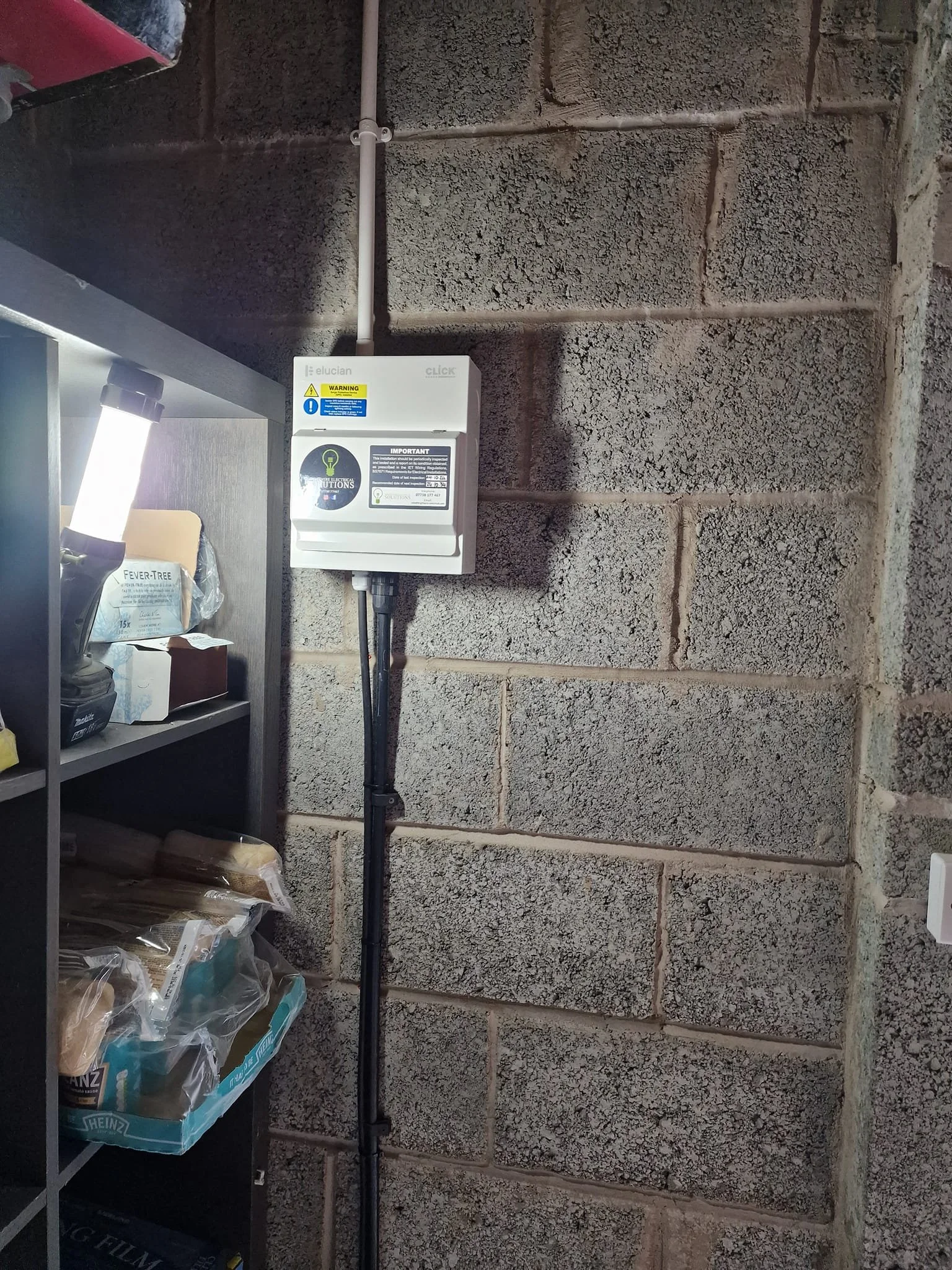A white electrical or utility box mounted on a cinder block wall in a storage or utility room, with black conduit pipes running down to the floor and shelves with assorted items including a box of Heinz baked beans and a box of face masks.