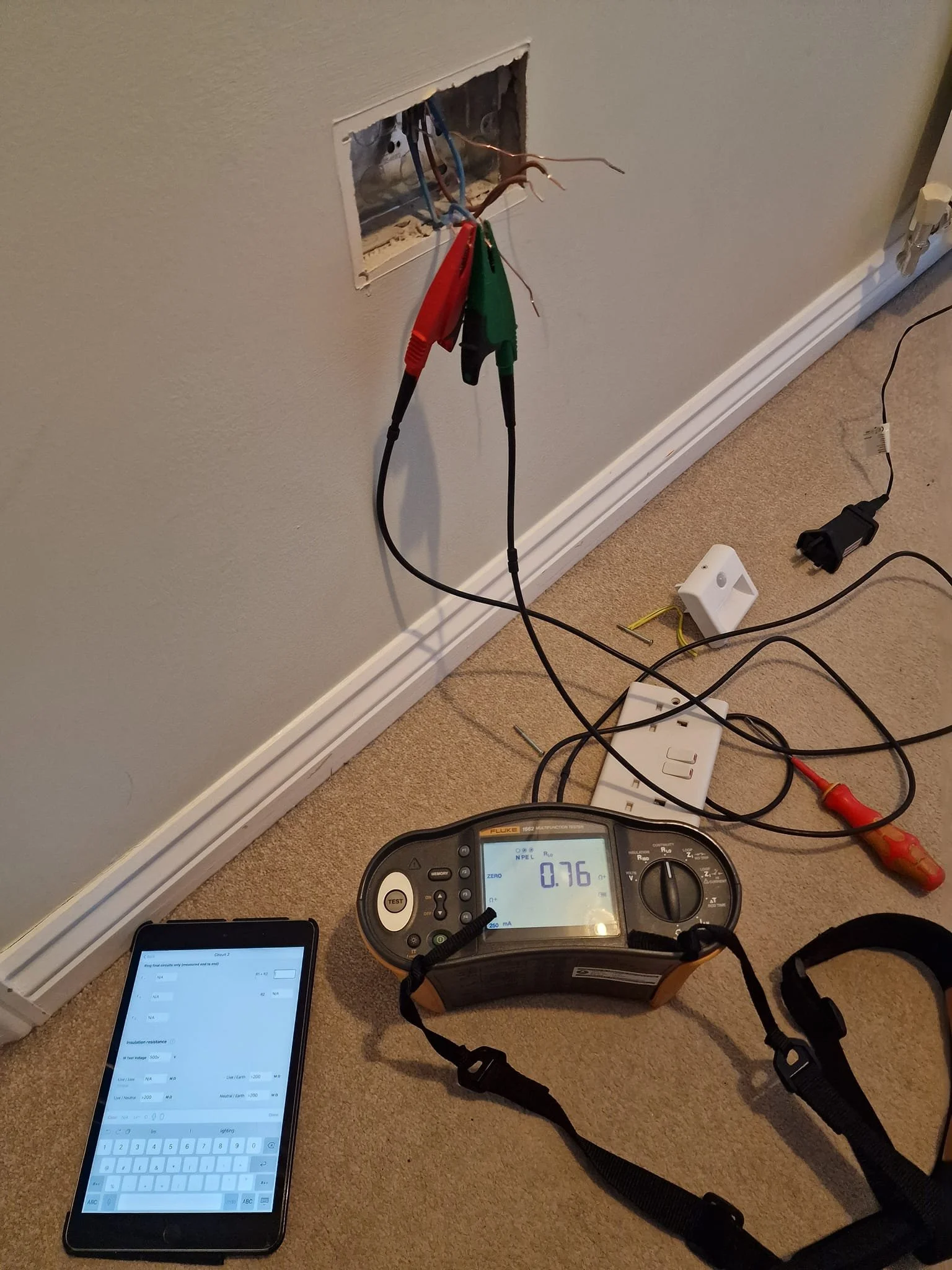 An electrical tester measuring 0.76 near a wall outlet with a partially open cover, testing wires with alligator clips, a device resembling a smartphone, a screwdriver, and a white plastic component on beige carpet.