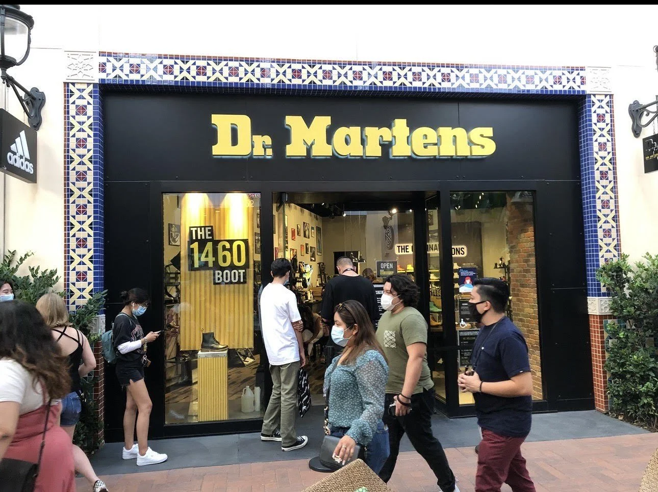 People walking outside a Dr. Martens store, with some waiting in line, wearing face masks, and the store's black facade with yellow signage and decorative tile border.