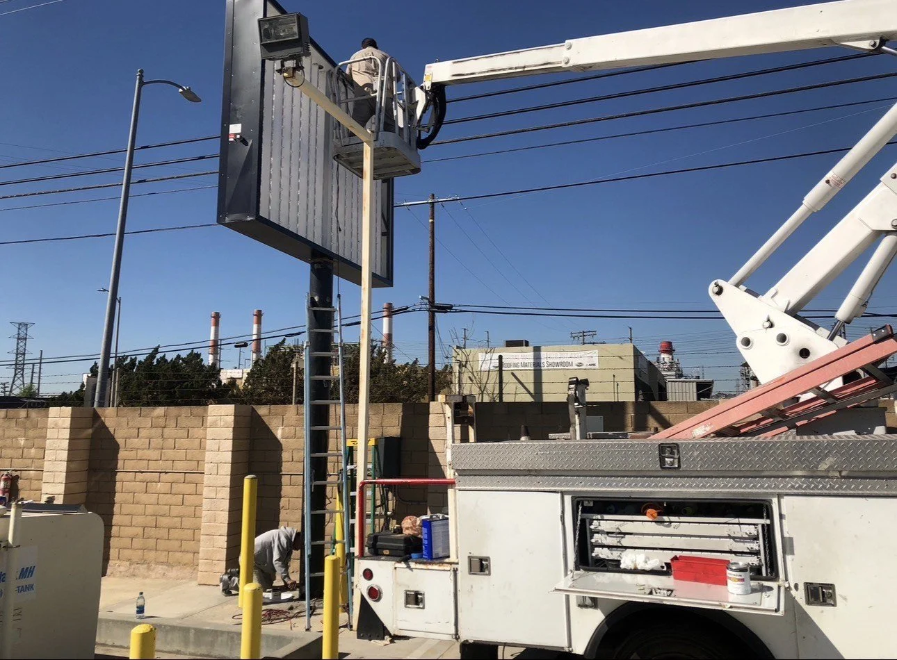 A worker on a bucket lift installing or repairing an electronic billboard at a street corner, with a brick wall, utility poles, and industrial buildings in the background.