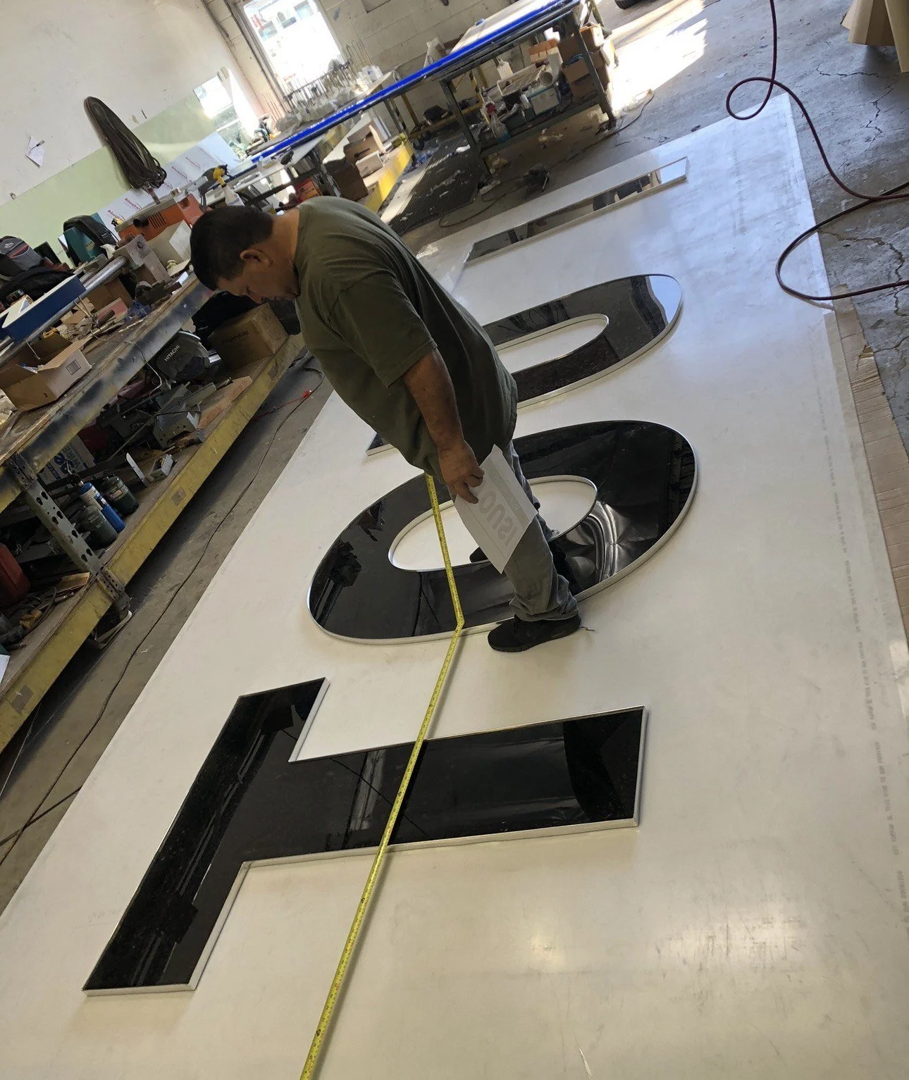 Man measuring a large white and black three-dimensional sign with cutout letters in a workshop.