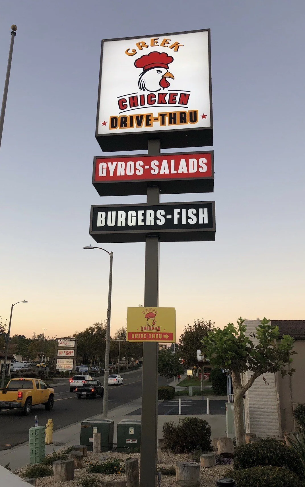 Sign for Greek Chicken Drive-Thru restaurant with separate signs for Gyros-Salads and Burgers-Fish, and a smaller sign with an arrow for the drive-thru. The signs are outdoors near a street with cars and trees.
