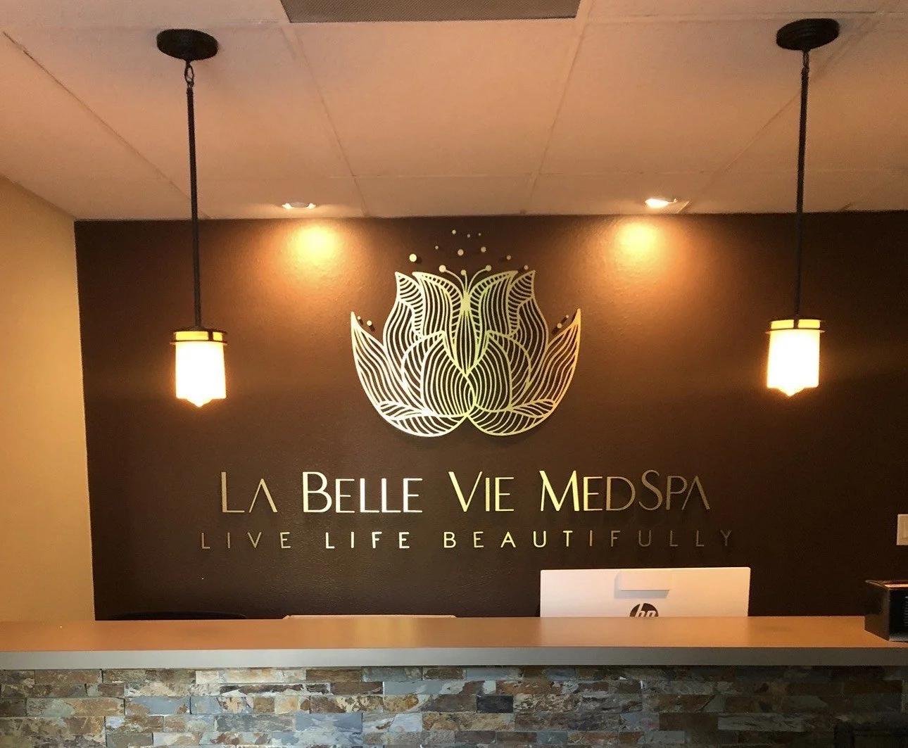 Reception area of La Belle Vie Med Spa with a black wall featuring a lotus flower logo, the spa's name, and the slogan 'Live Life Beautifully,' illuminated by two hanging pendant lights.