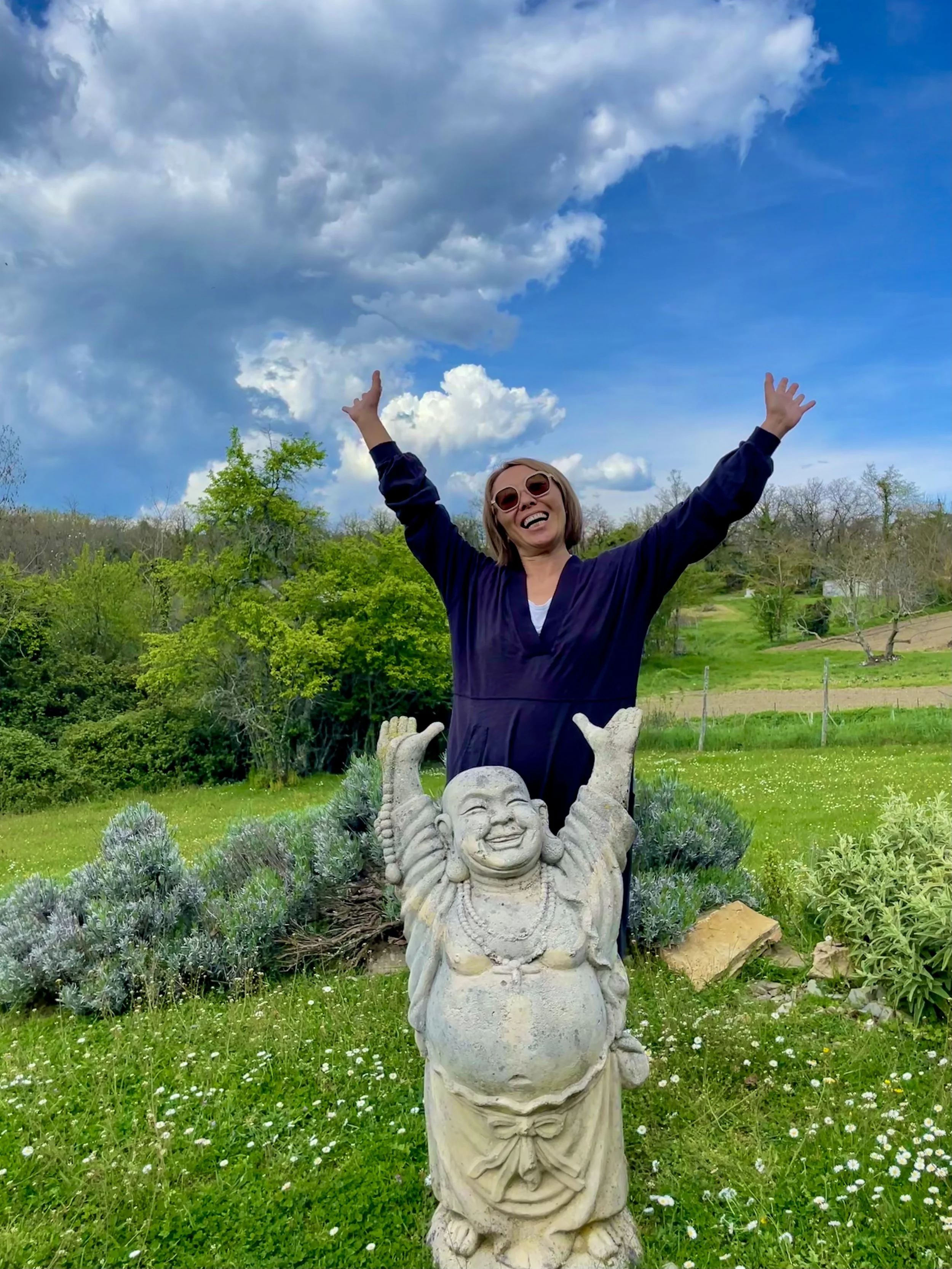 A woman wearing sunglasses and a dark jacket is standing outdoors behind a laughing smiling sculpture with arms raised. The background features a green landscape, trees, and a partly cloudy blue sky.