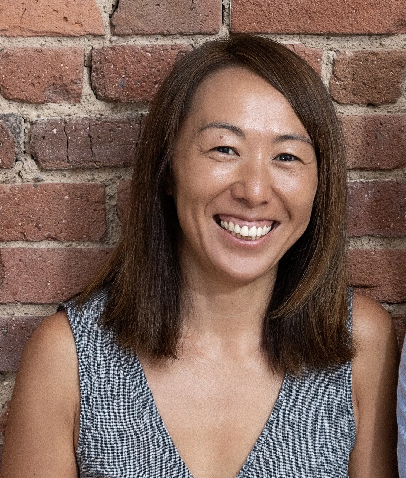 A woman with shoulder-length brown hair, smiling, wearing a gray sleeveless top, standing in front of a brick wall.