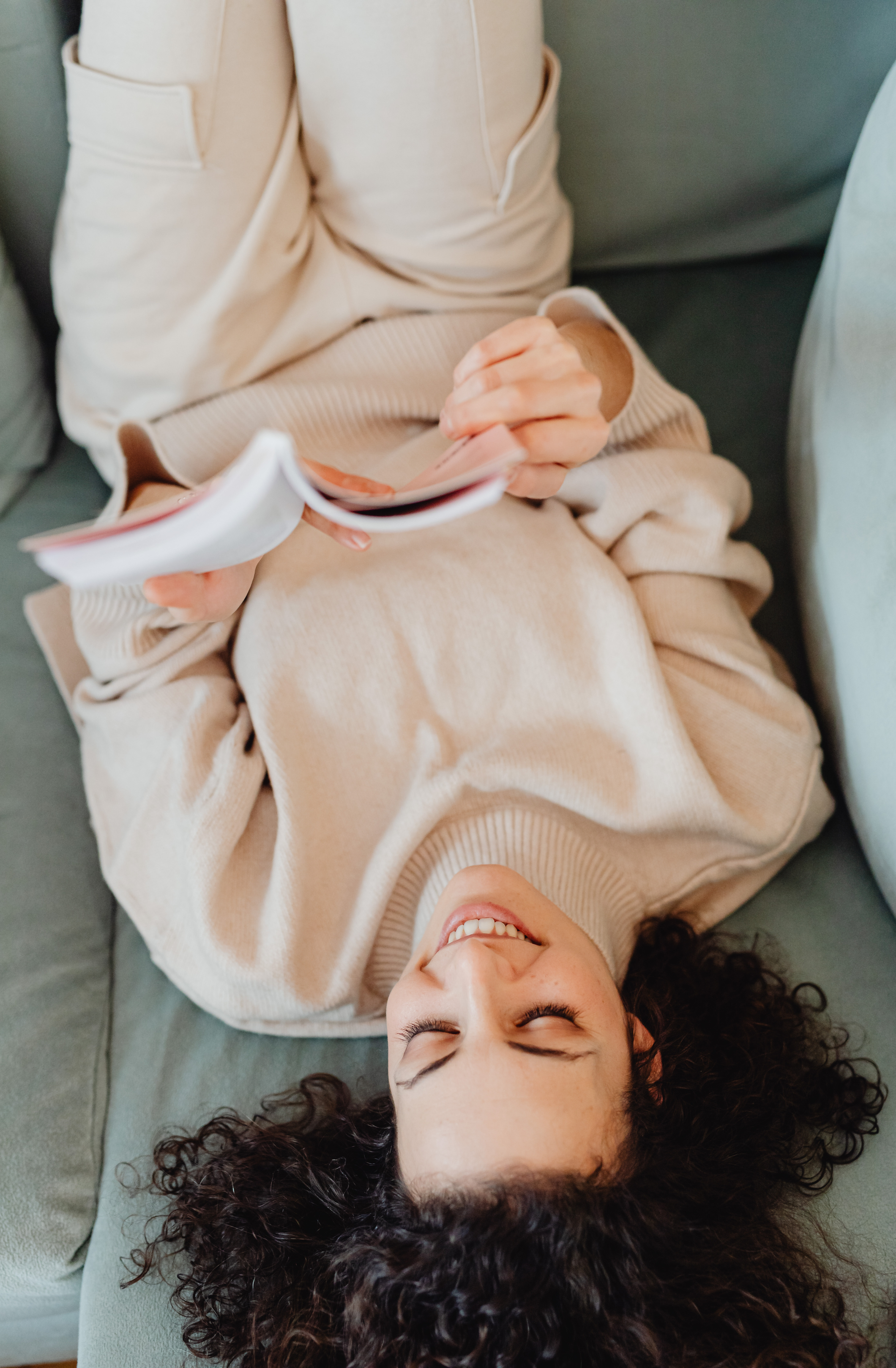 A young woman with curly dark hair lying on a couch, smiling with her eyes closed, and holding a magazine.