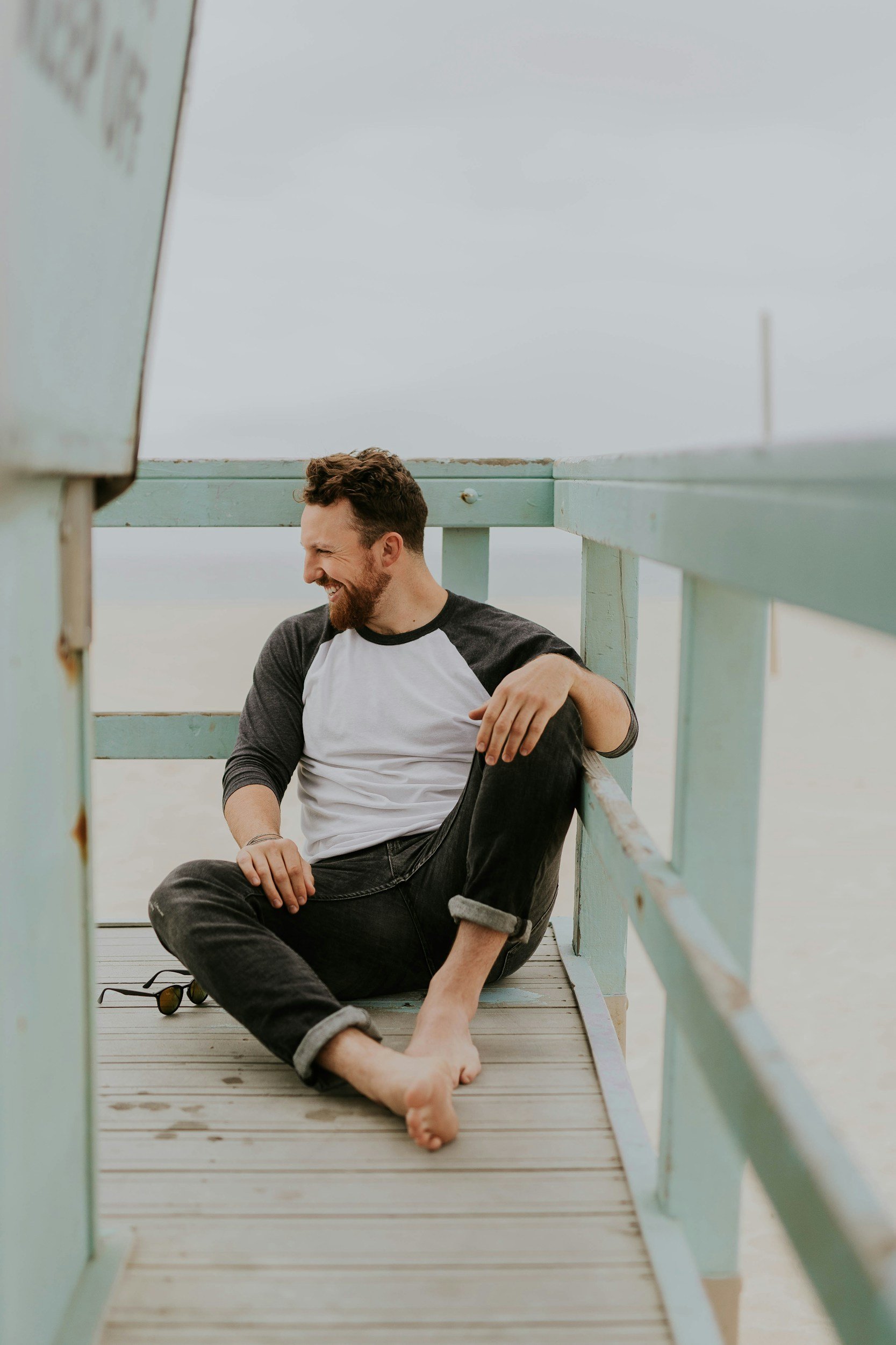 A man with a beard and curly hair, dressed in a white and black baseball tee and rolled-up jeans, sitting on a wooden pier, smiling and looking to the side.