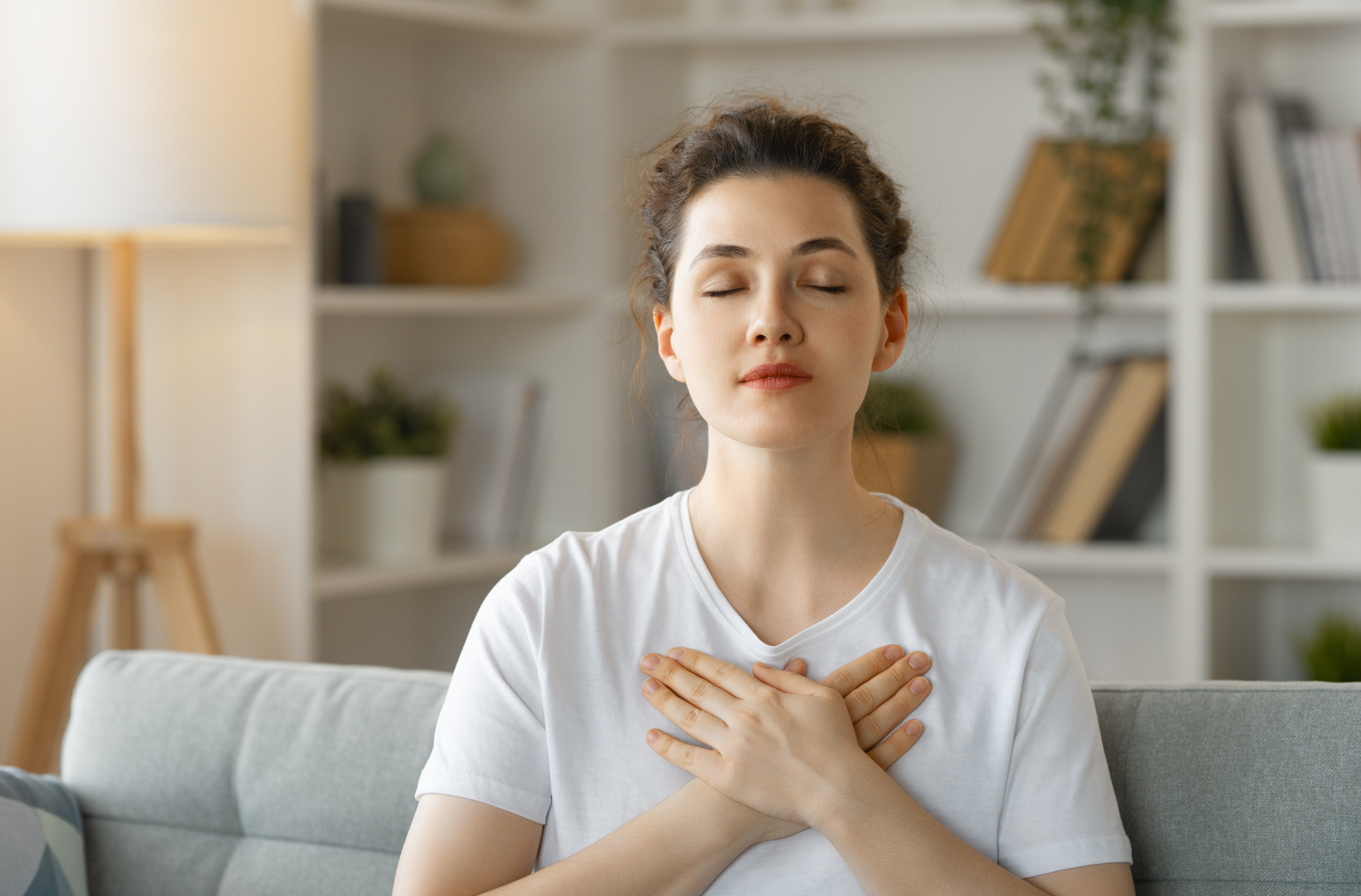 A woman sitting on a sofa with her eyes closed and hands on her chest, appearing to practice mindfulness or meditation in a cozy living room.