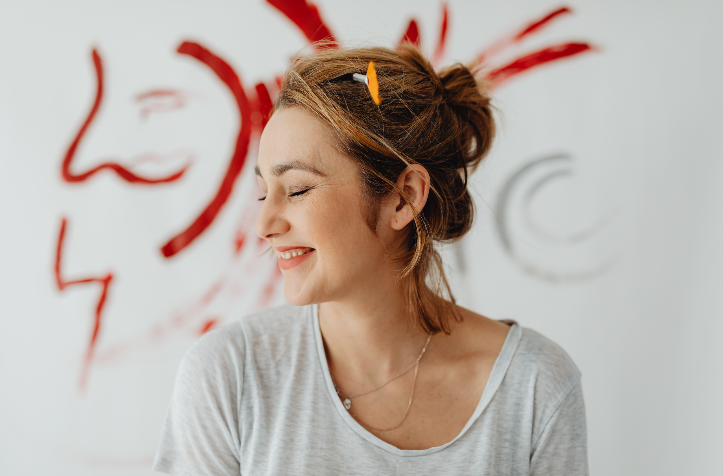 Woman with a smile, closed eyes, and messy hair, wearing a light gray t-shirt and a necklace, in front of a wall with red and gray artwork.