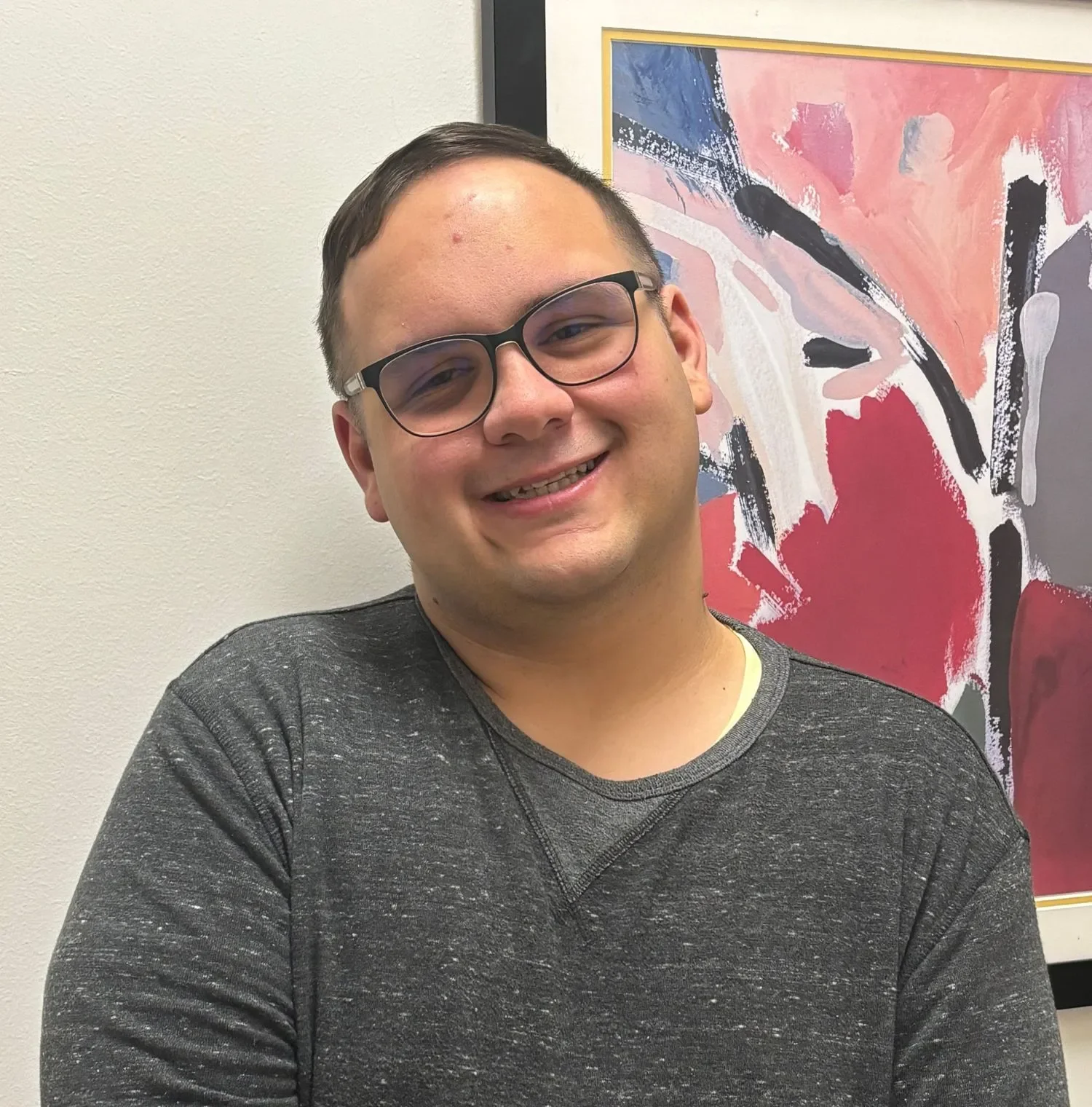 A smiling young man with glasses wearing a dark gray sweater, standing in front of an abstract colorful painting on a wall.