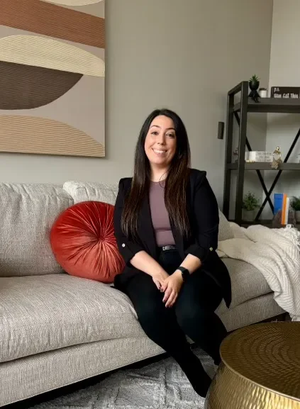 A woman sitting on a gray couch in a living room smiling at the camera. She has long dark hair and is wearing a black blazer over a gray top. There is a red round pillow beside her and a black shelf with decor and books behind her.