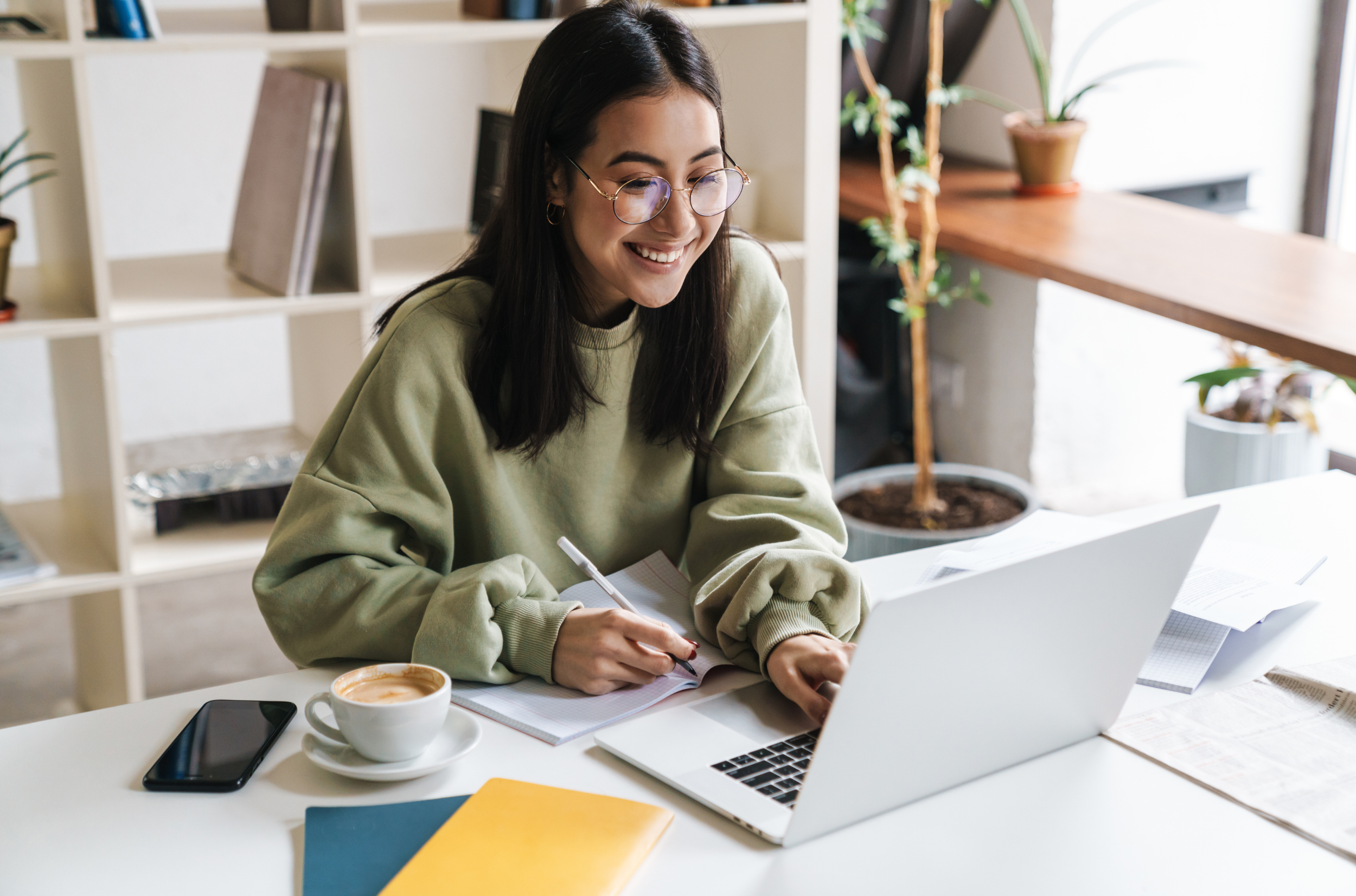 Smiling young woman with glasses working on laptop at a white desk in a cozy, well-lit room.