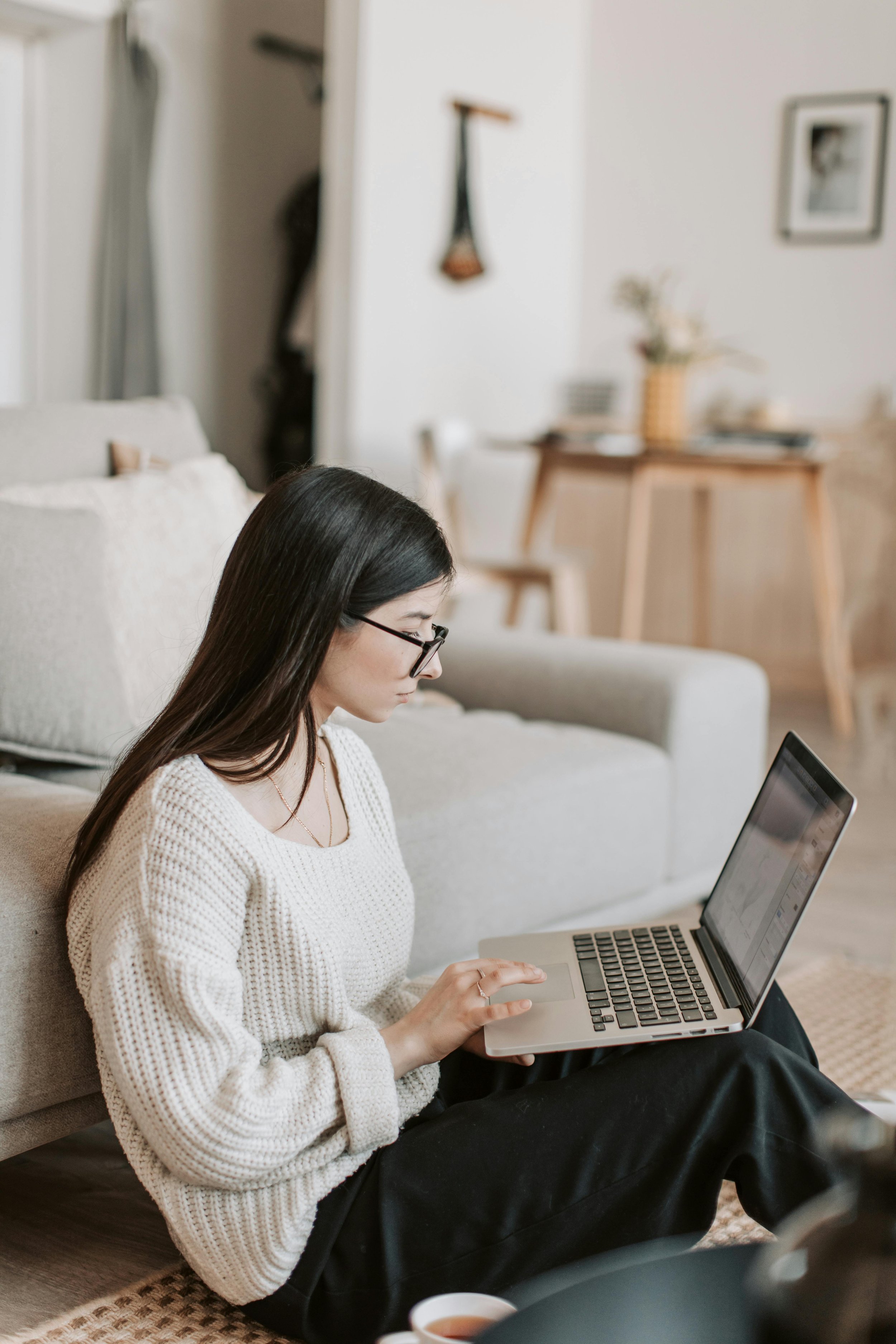 A woman with long dark hair and glasses sitting on the floor working on a laptop in a cozy living room.