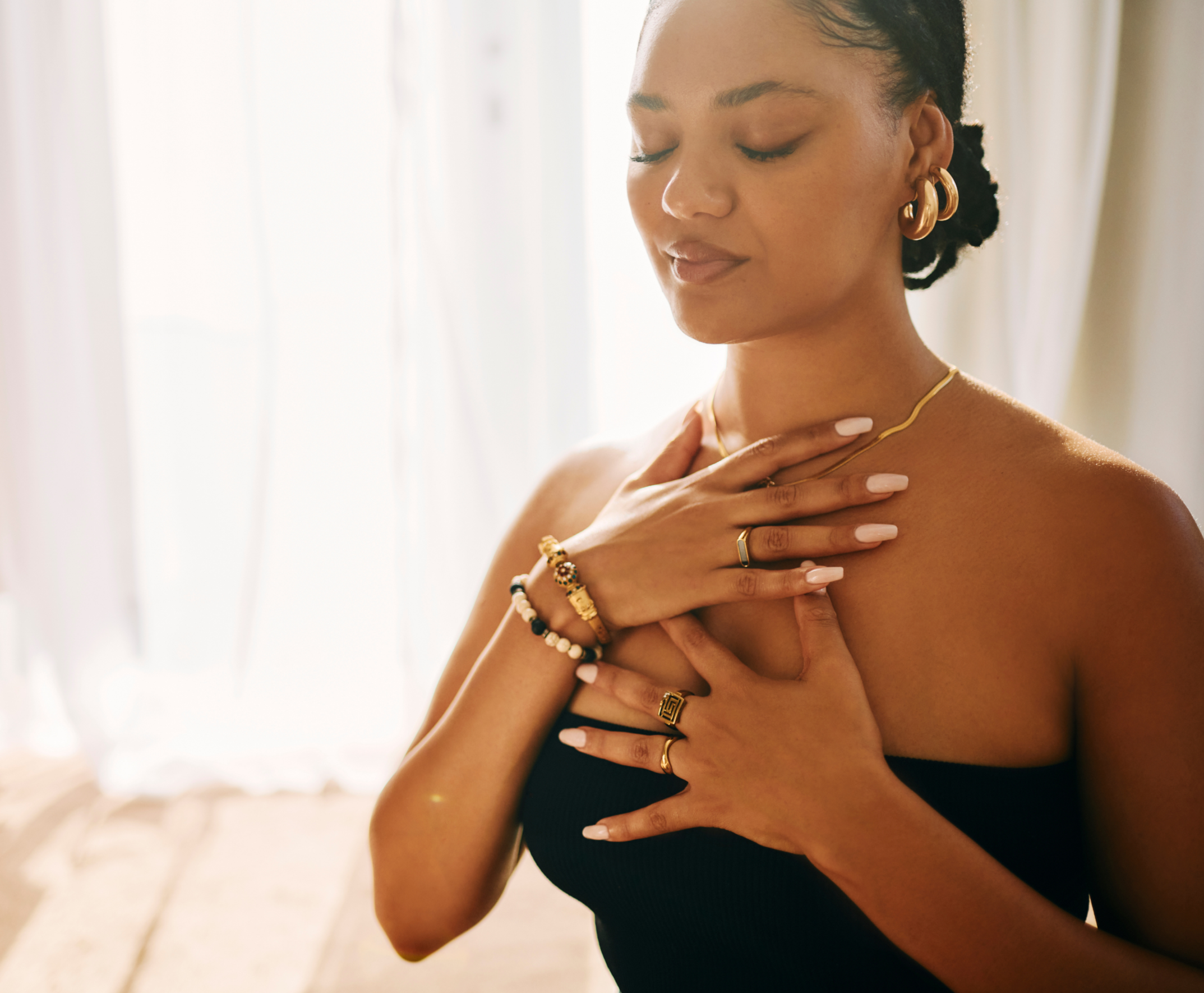 A woman with dark hair in a low bun, wearing gold jewelry and a black strapless top, stands with her eyes closed, hands on her chest, in a softly lit room with white curtains.