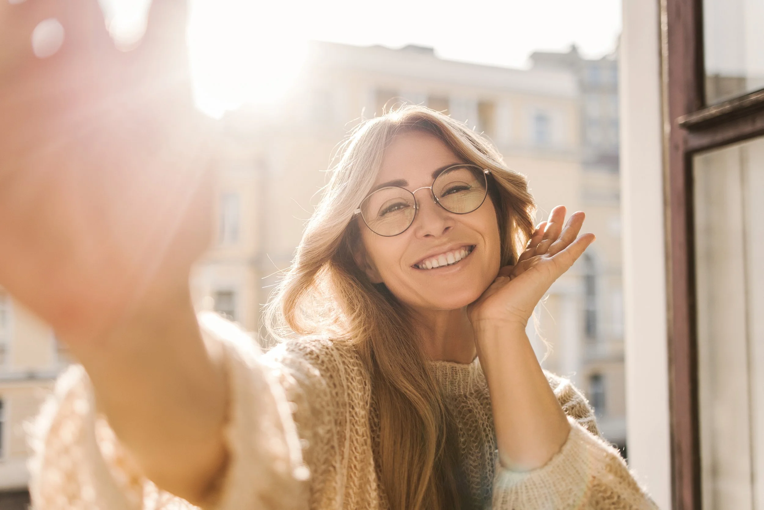 A woman with glasses smiling and waving, taking a selfie by a window with sunlight in the background.