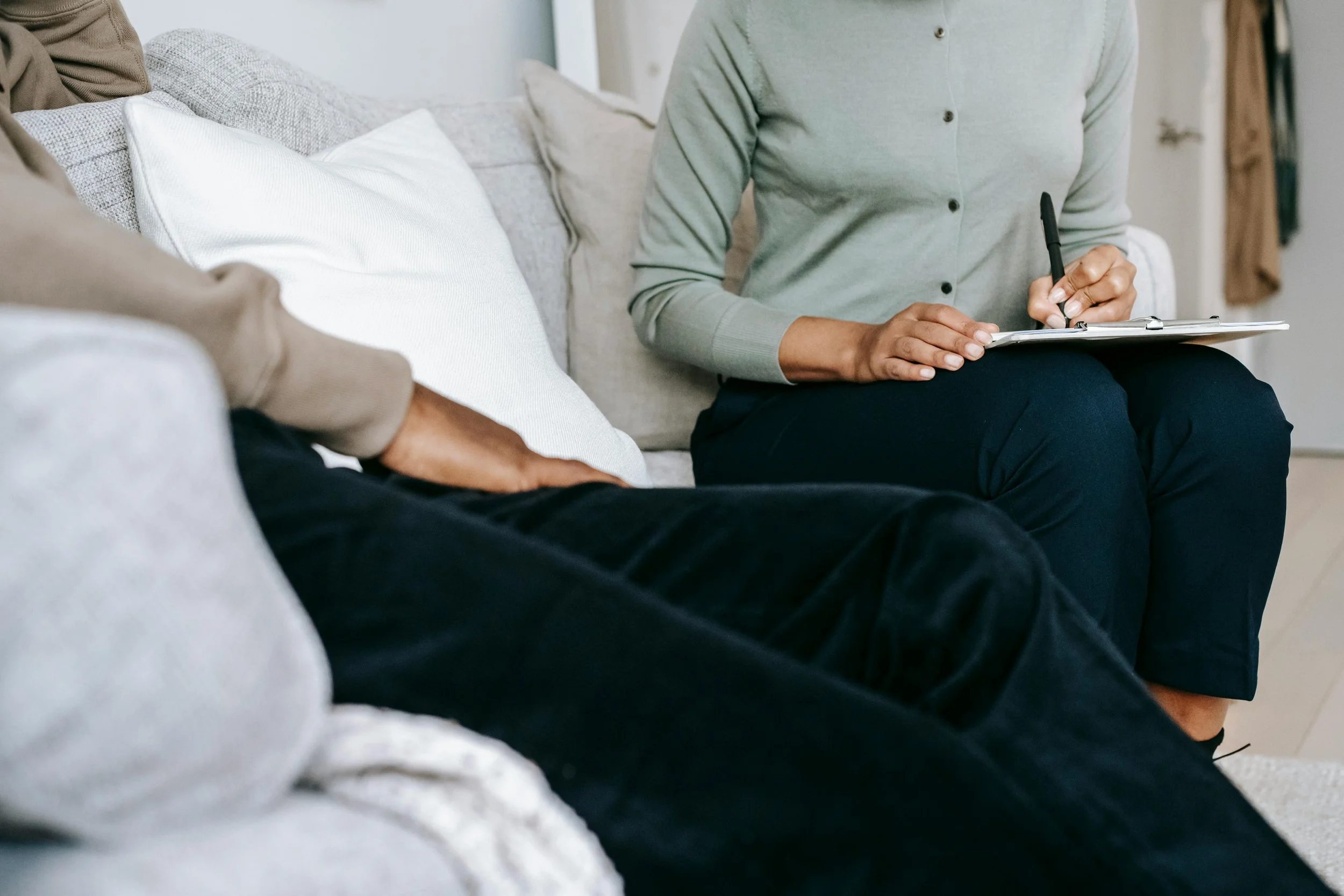 A person sitting on a couch with hands resting on their lap, wearing a beige shirt and black pants, while another person is sitting next to them, taking notes or filling out a form.