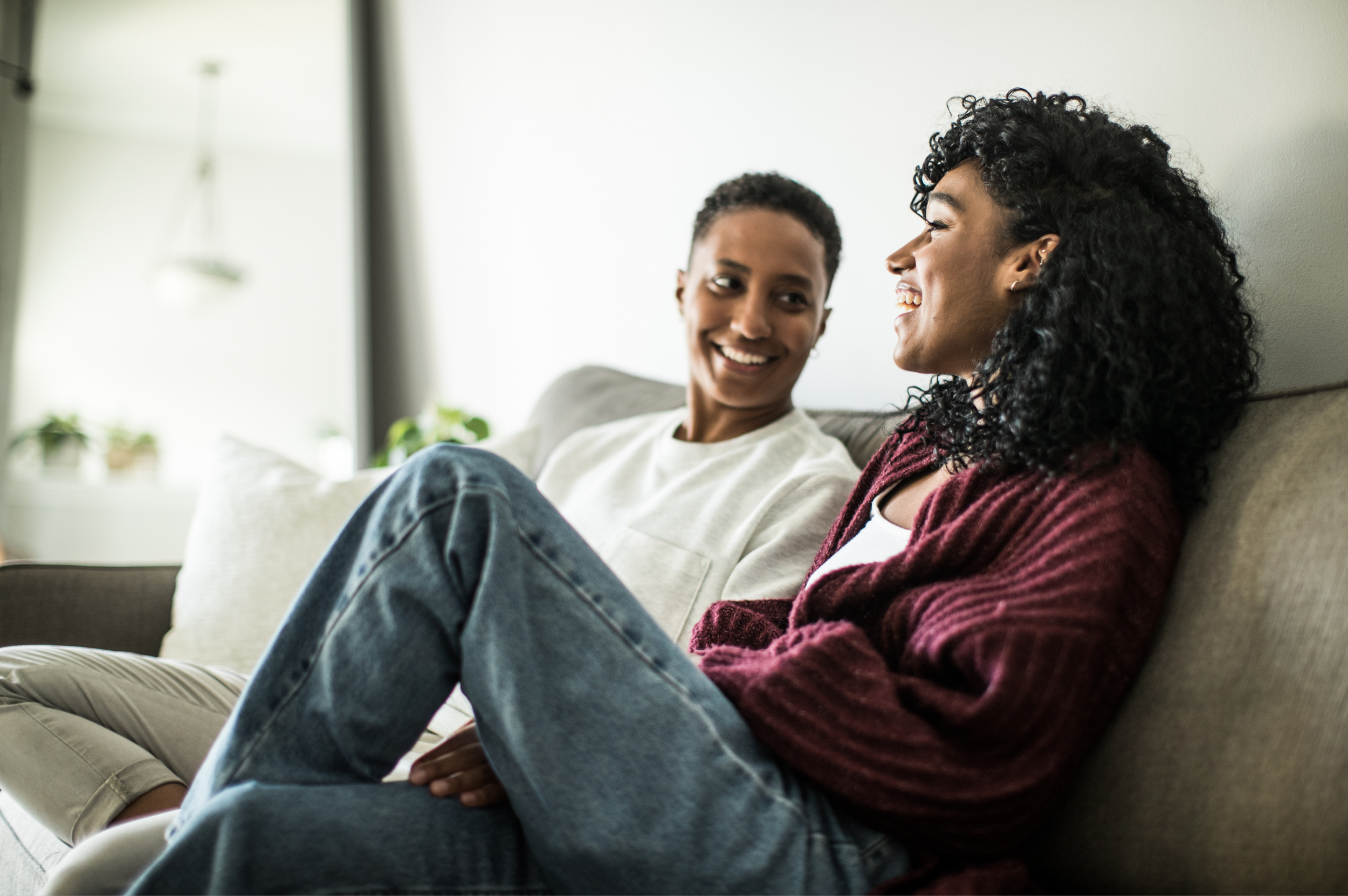 Two women sitting on a beige couch and smiling at each other in a living room.