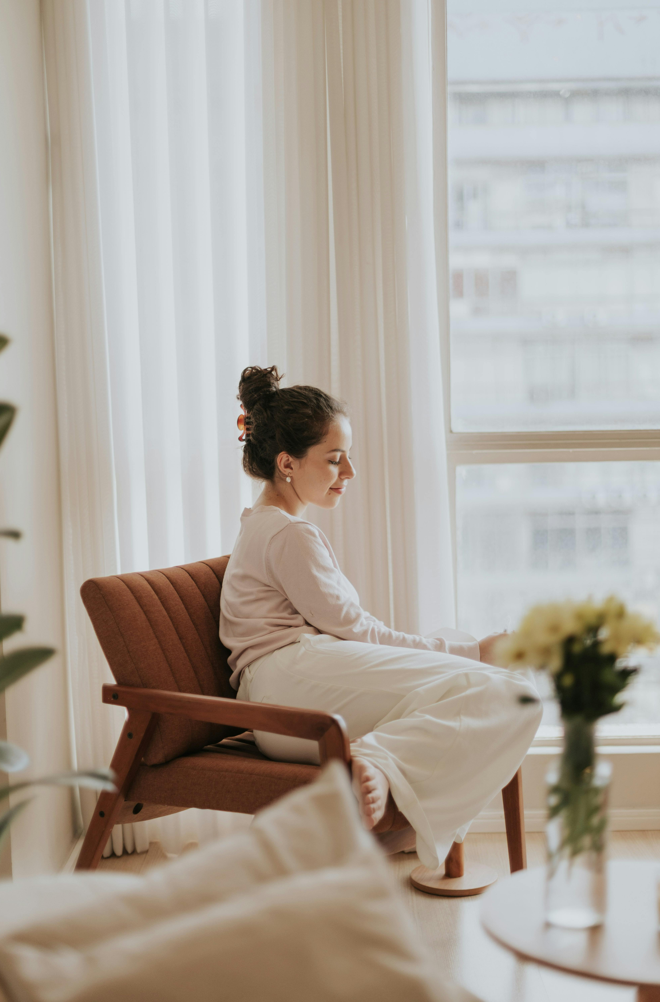 A woman sitting on a brown armchair by a large window with white curtains, looking down with a peaceful expression.