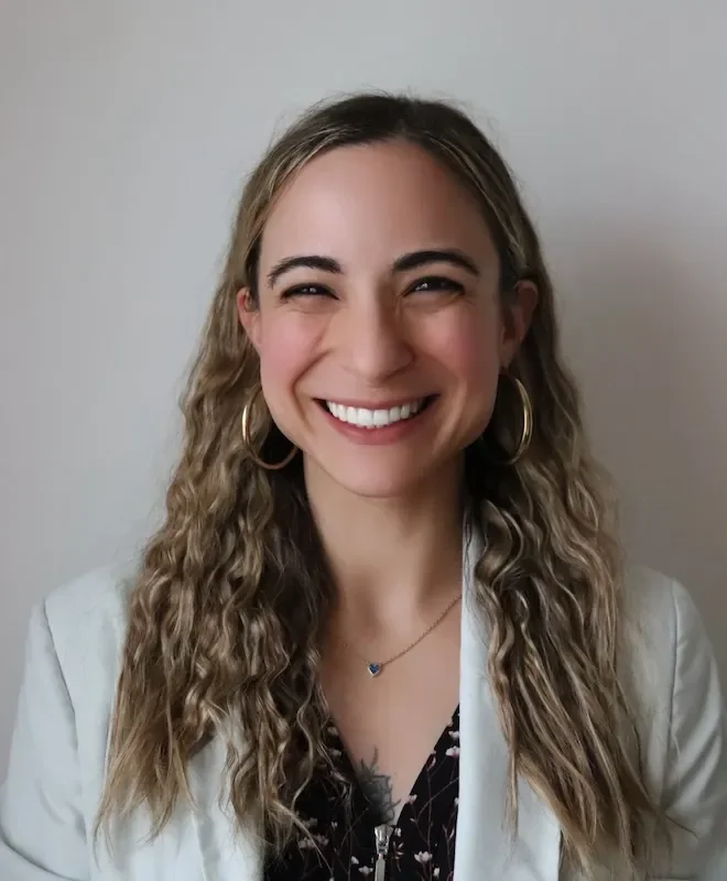 A smiling woman with curly blonde hair, wearing hoop earrings, a white blazer, and a black floral top, standing against a plain wall.