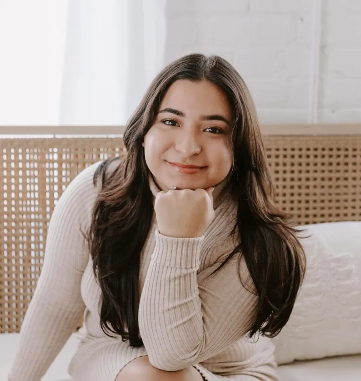 A woman with long dark hair sitting on a bed, smiling, resting her chin on her hand in a cozy room with a woven headboard and white brick wall background.