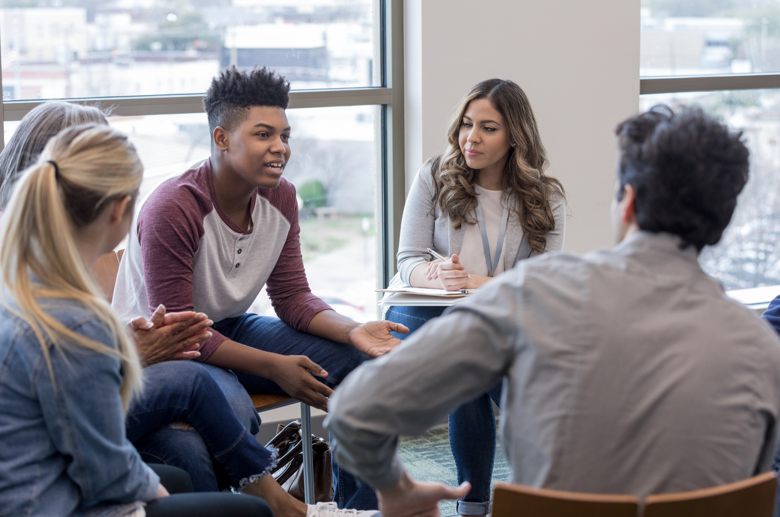 Group of young people engaged in a discussion in a bright room with large windows.