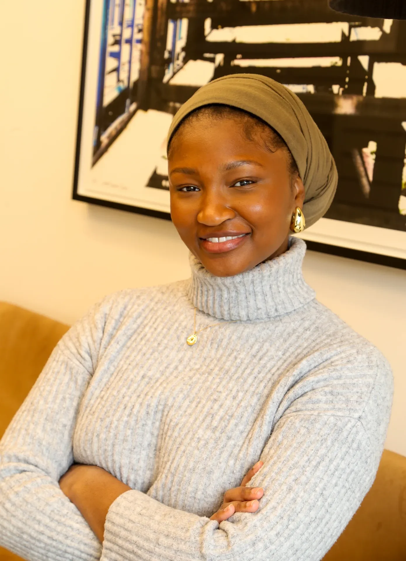 A smiling woman with a tan headscarf, gold earrings, and a beige turtleneck sweater, standing with arms crossed in front of a wall with a black and white framed photograph.